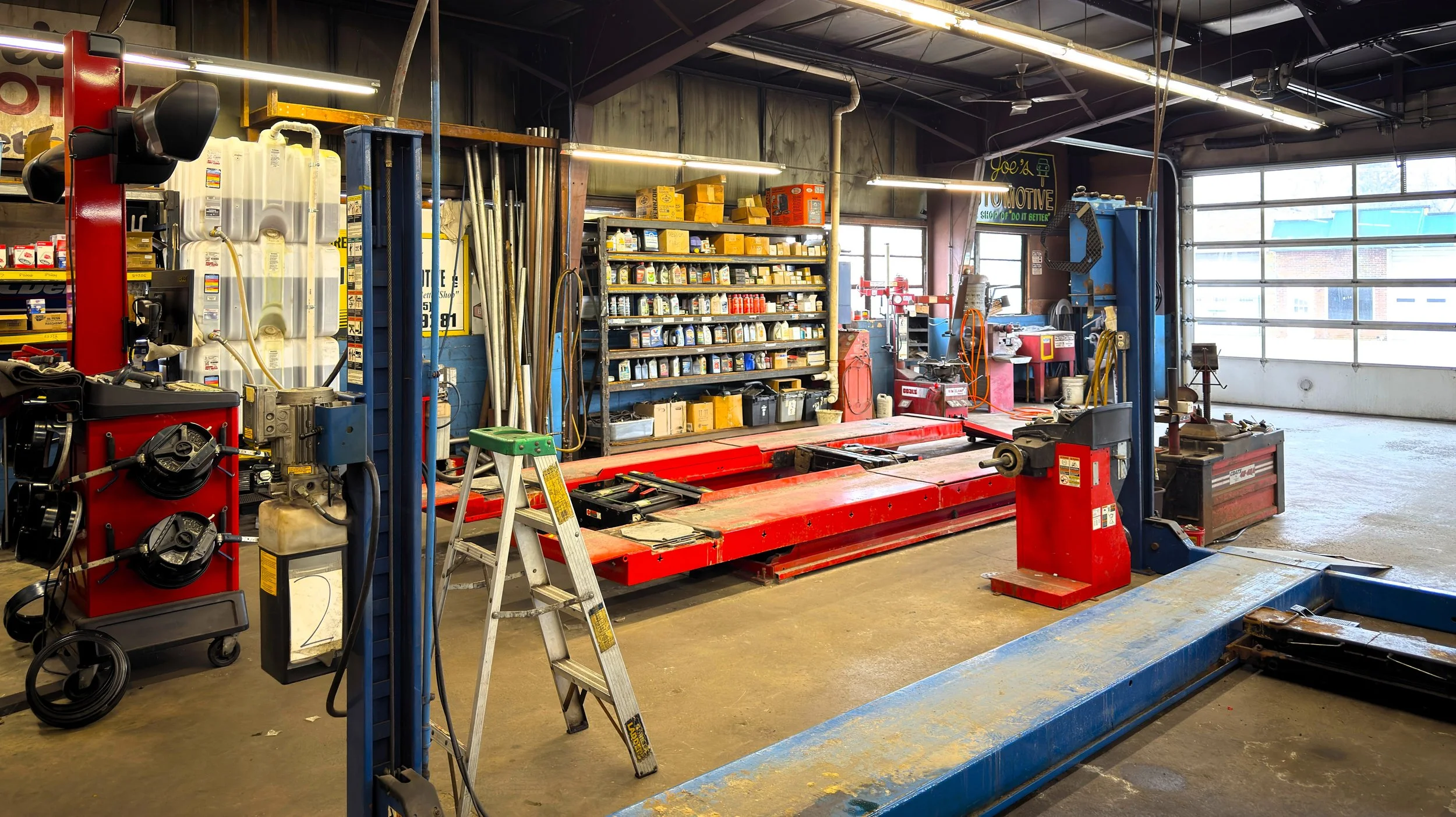 Inside an auto repair shop with a vehicle lift, shelves of car parts, a step ladder, and various tools and equipment.