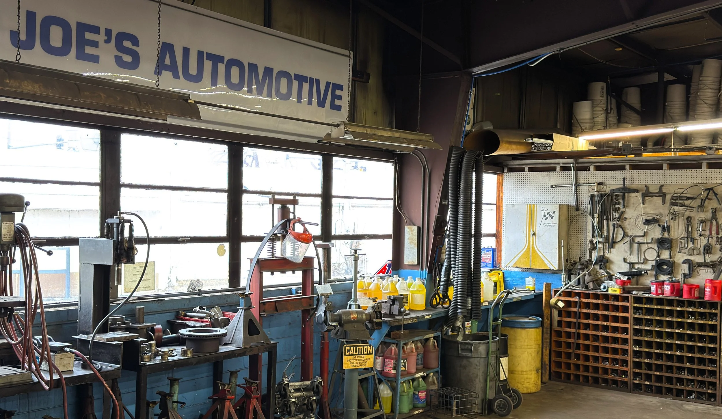 Inside an auto repair workshop with tools and equipment, a workbench with various mechanical parts, yellow engine oil containers, a pegboard with hand tools, and a sign reading 'Joe's Automotive'.