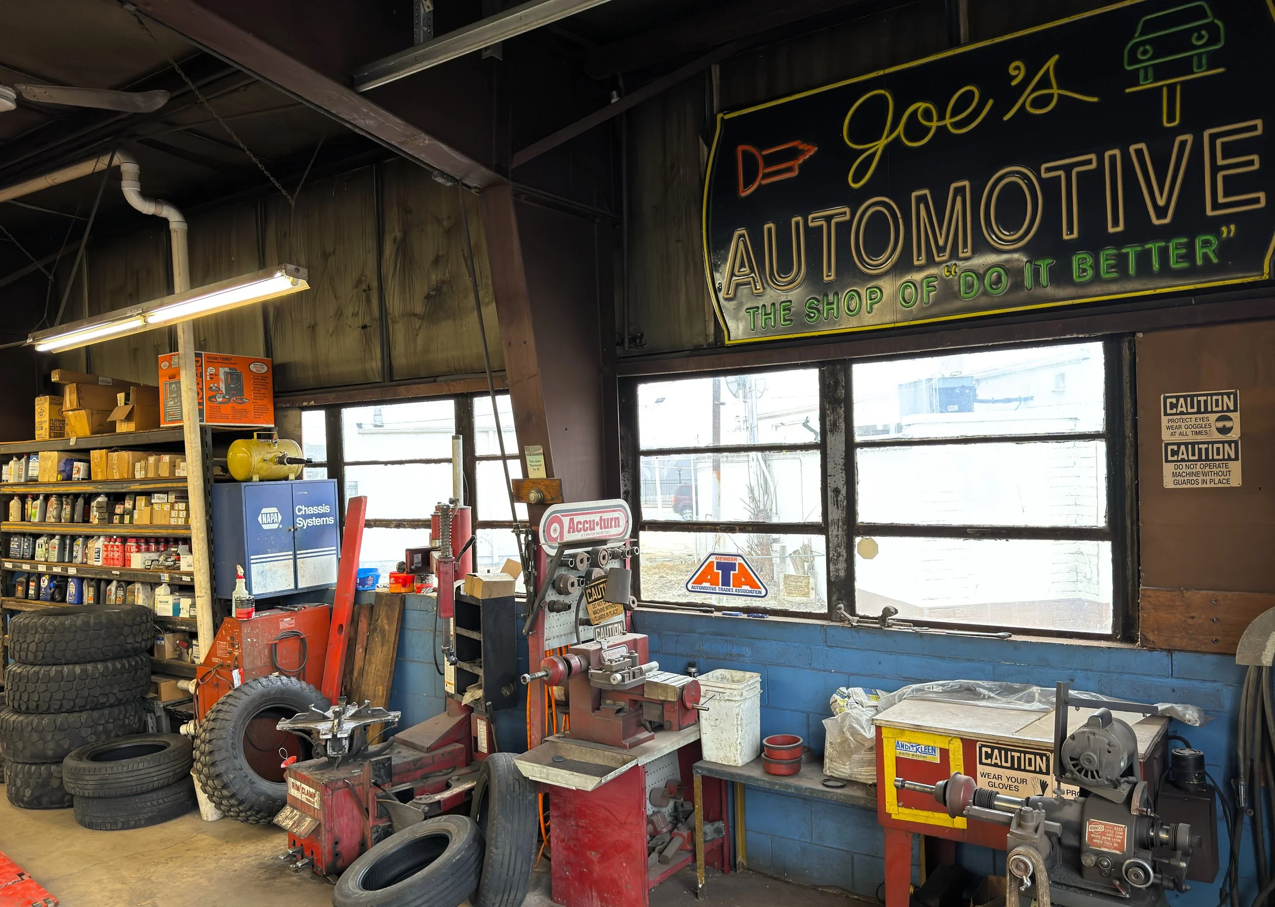 Inside an automotive repair shop with tires, tools, and equipment. A large window, shelves with car parts, and a neon sign that reads 'Joe's Automotive, the shop of 'do it better.'