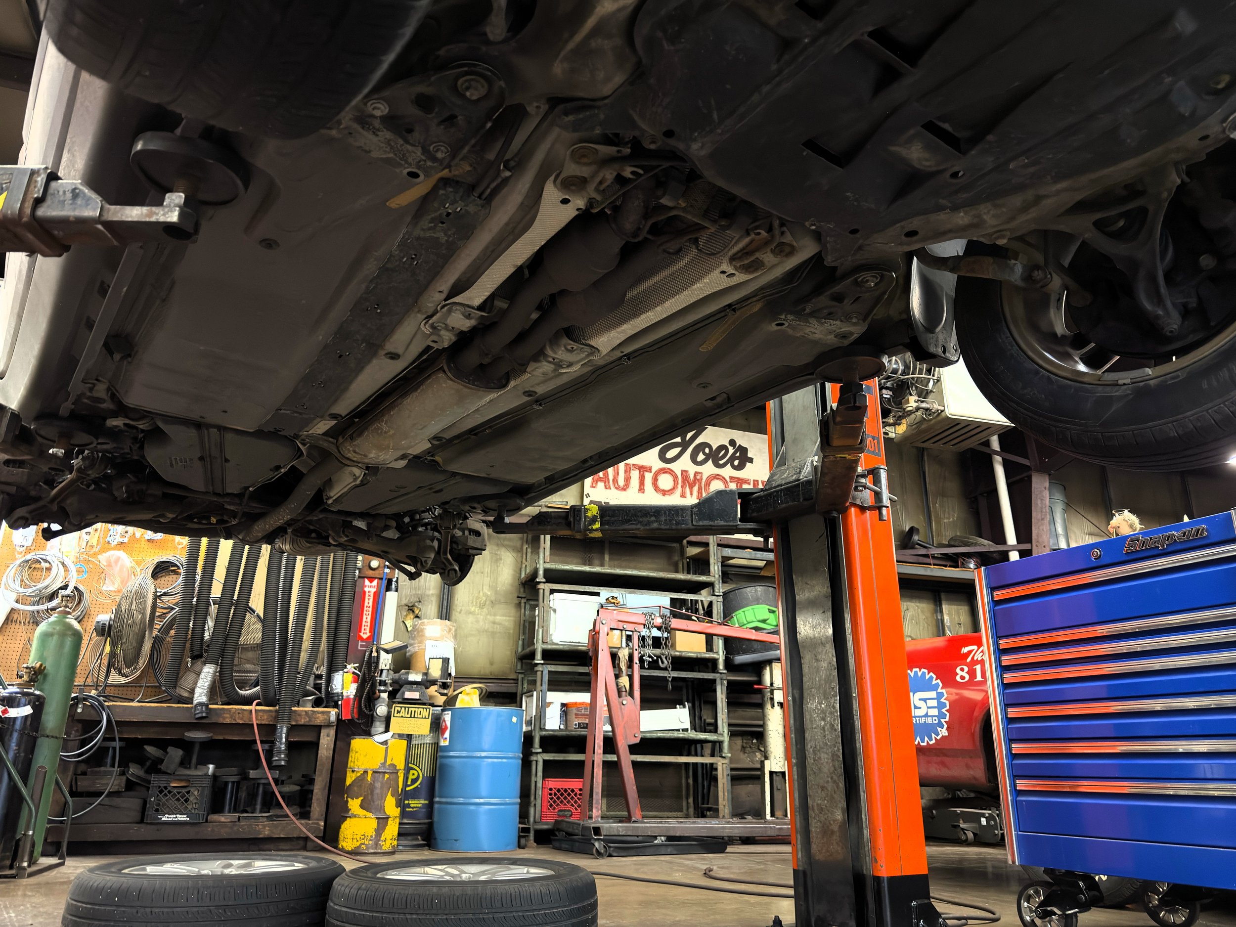 Underneath view of a car lifted in a mechanic's garage with tools, equipment, and tires around.