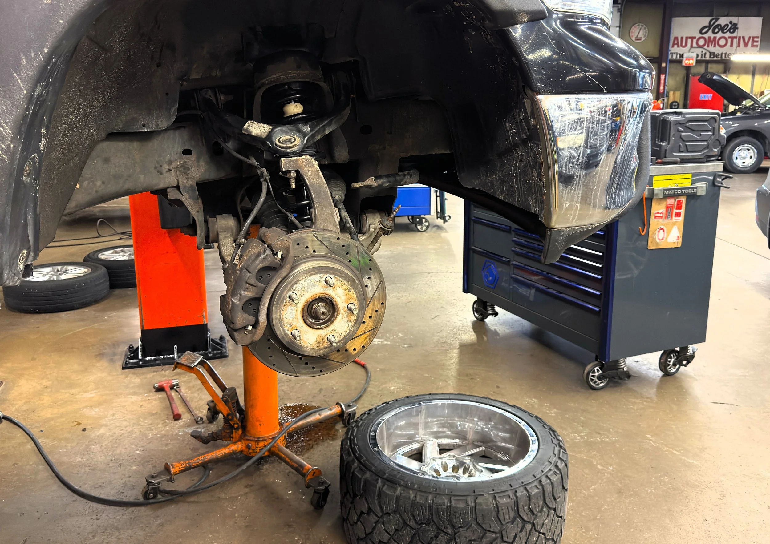 A vehicle is lifted on a hydraulic hoist inside an automotive repair shop. The front wheel and brake assembly are removed, exposing the brake rotor and suspension components. A tire and rim are placed on the floor nearby, and various tools and equipment are in the background.