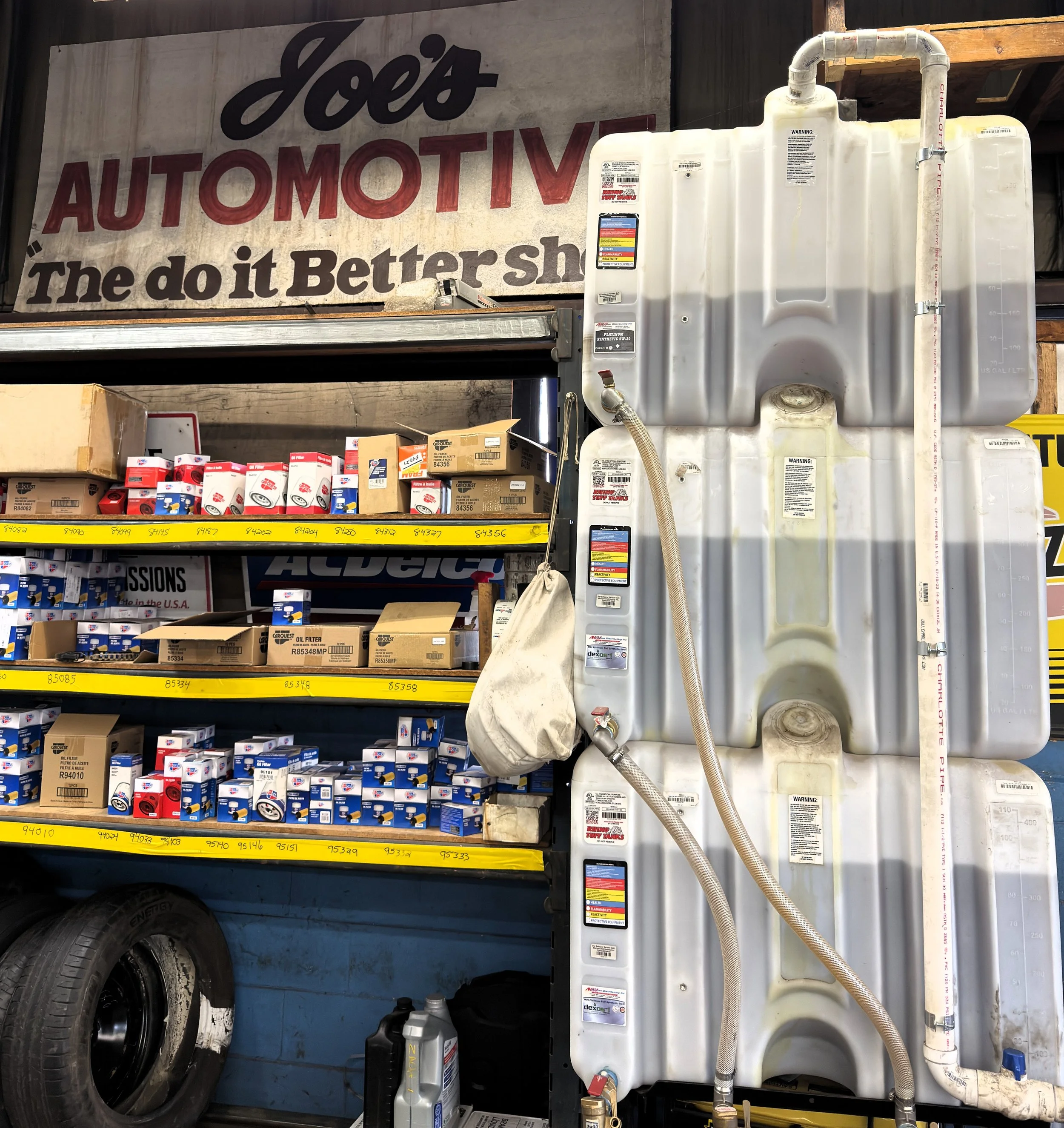 Auto parts store aisle with shelves stocked with car filters and boxes, tire on the bottom left, and large white water tanks with pipes on the right, and a sign reading 'Joe's Automotive' in the background.