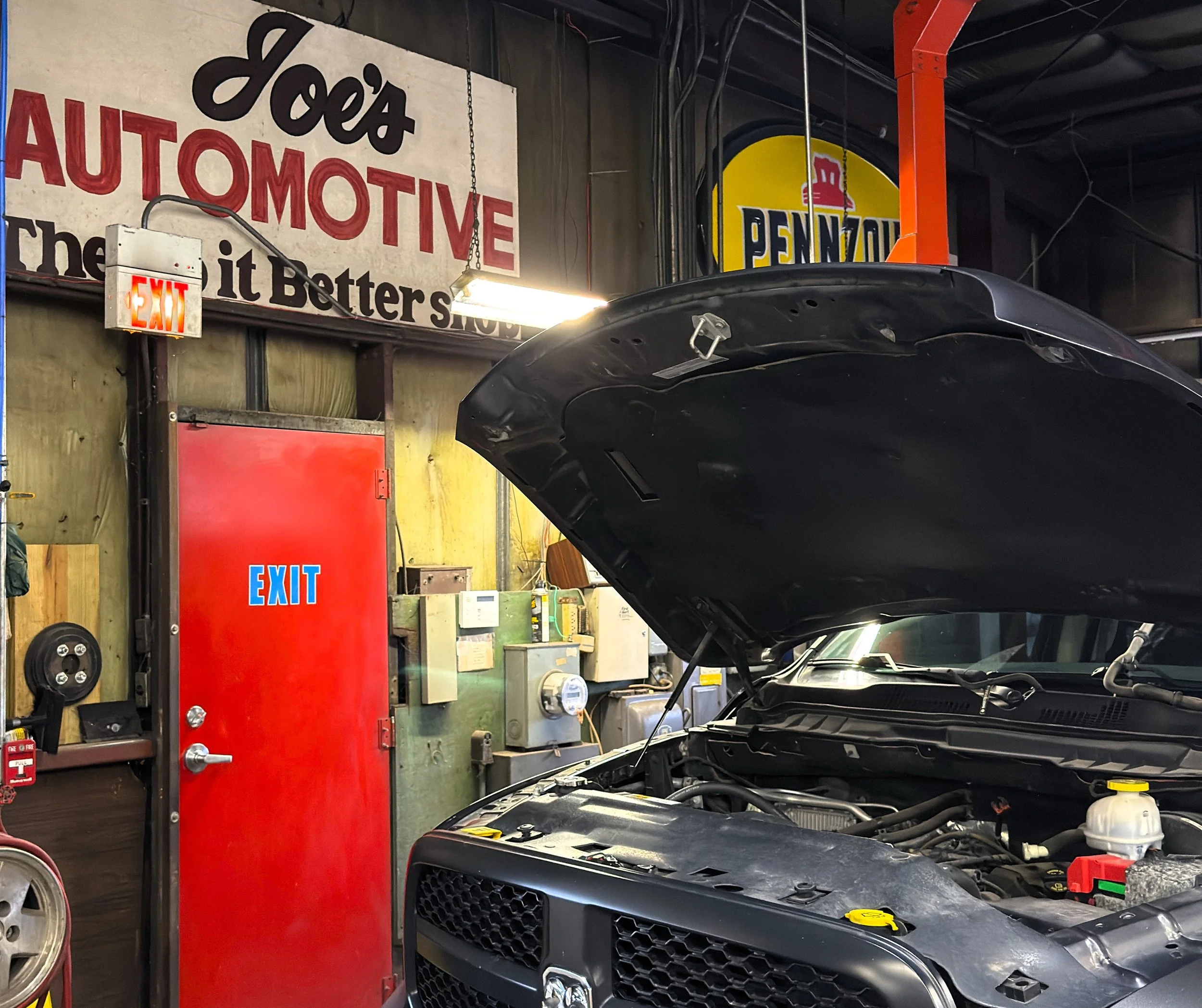 An open car hood in an automotive repair shop with a red exit door and various tools and equipment hanging on the wall. There are signs including 'Joe's Automotive' and 'Pennzoil' in the background.