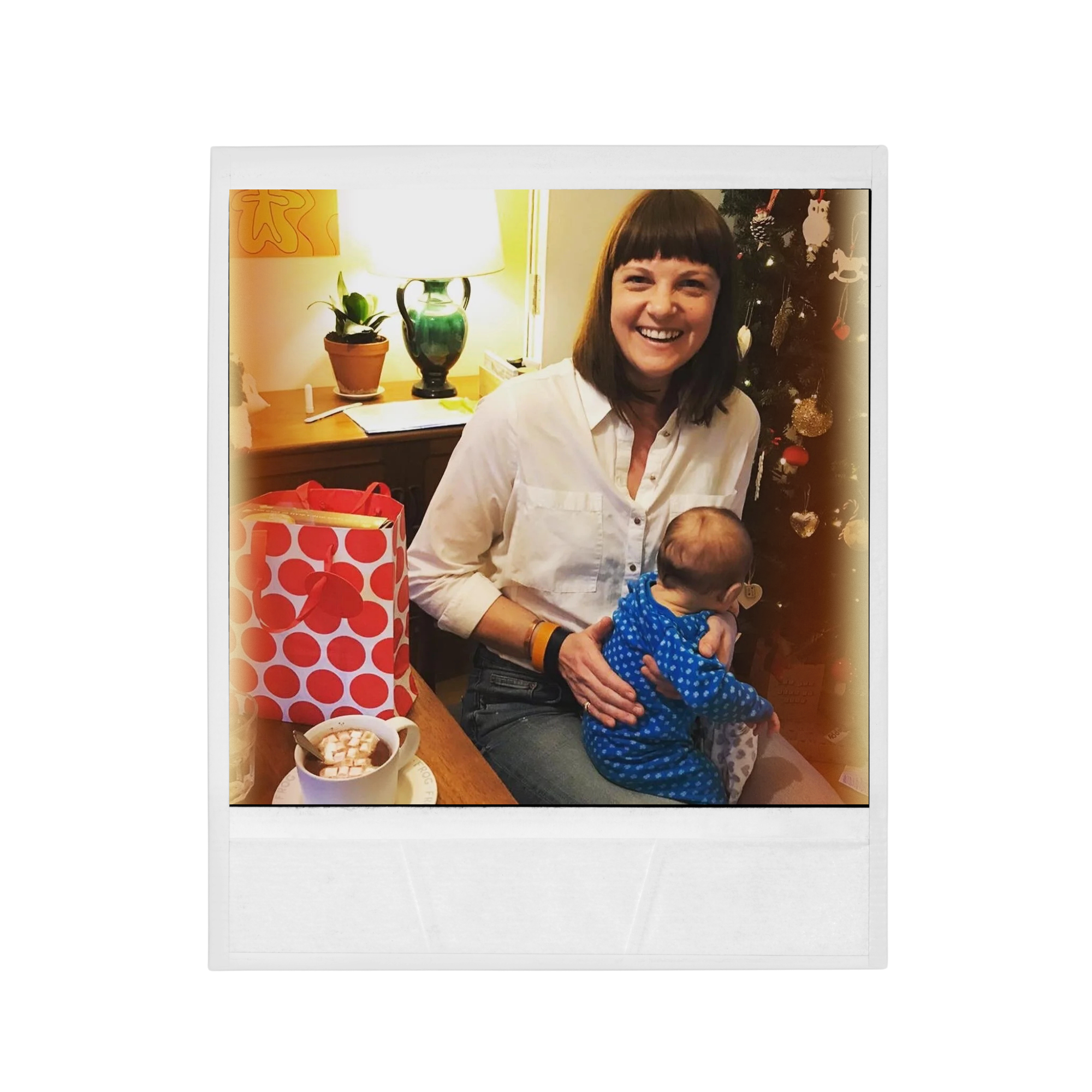 A smiling woman with shoulder-length brown hair wearing a white shirt holding a young child in a blue polka dot outfit in front of a decorated Christmas tree. The setting appears to be inside a home with a lamp, potted plant, and some papers on a wooden table.