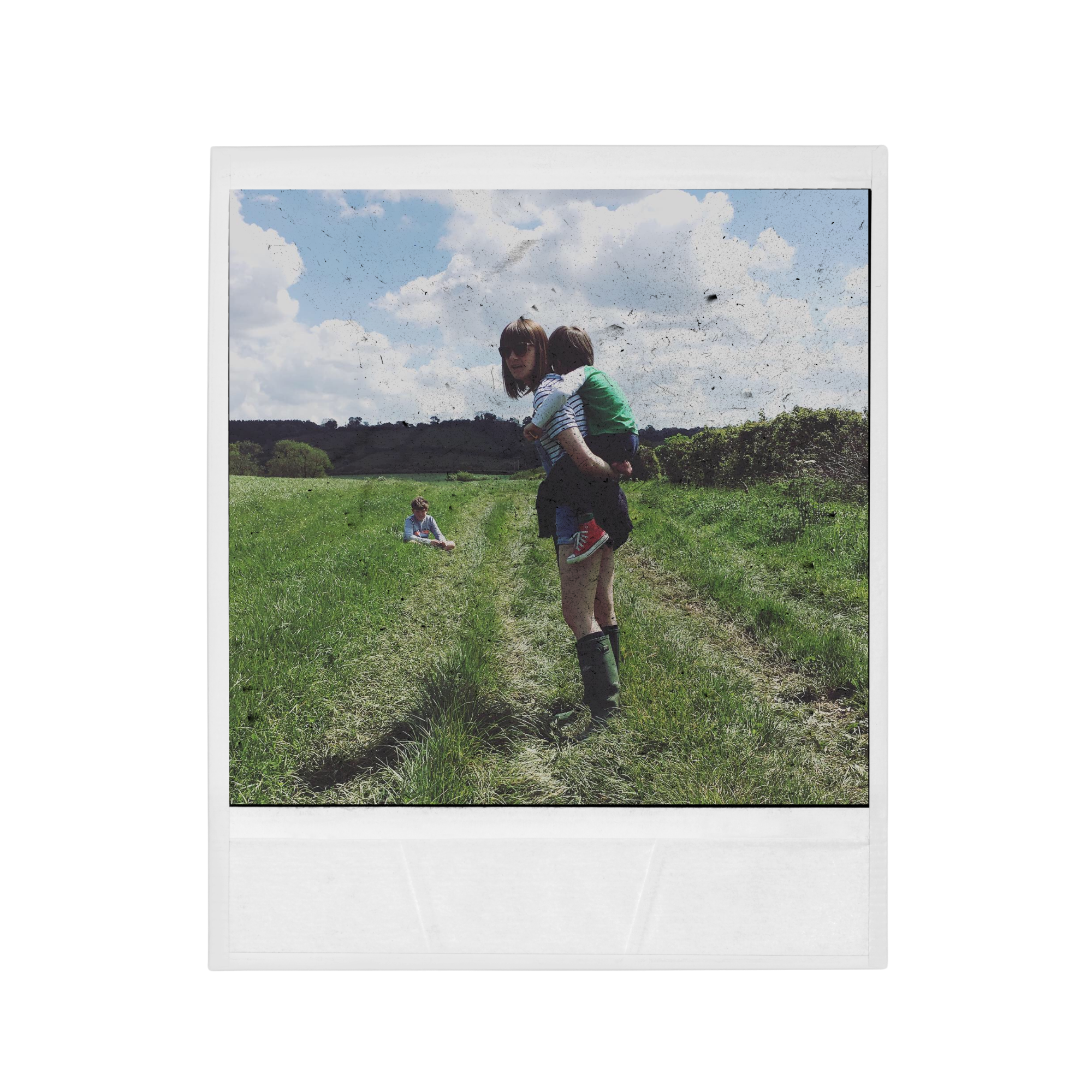 Picture of a polaroid photo featuring two kids playing outside on grassy fields with trees and hills in the background, under a partly cloudy sky.