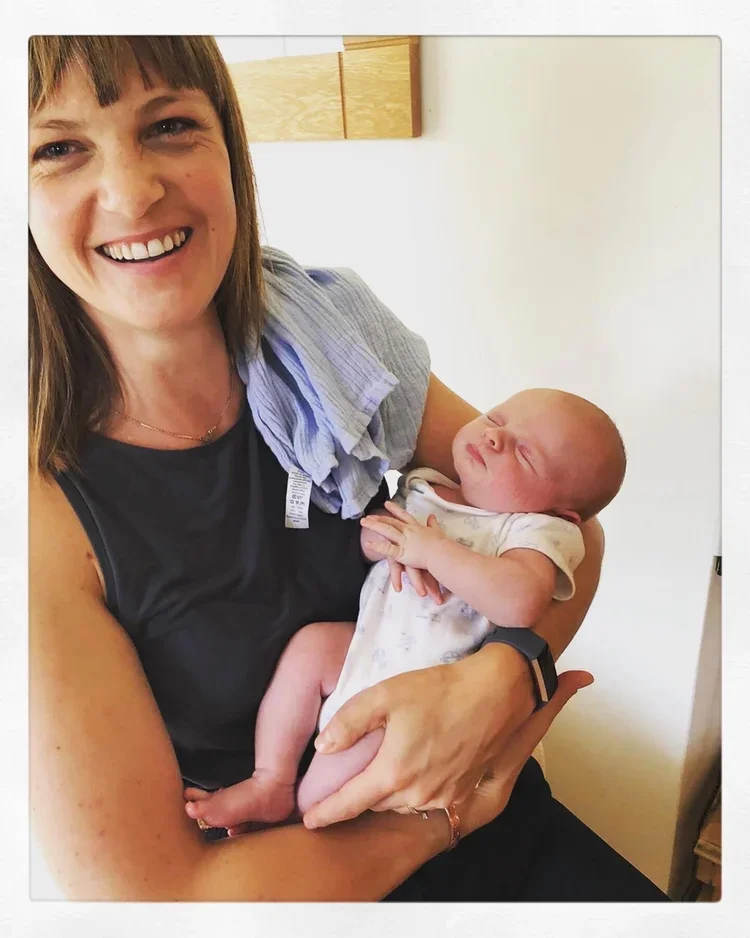 A woman holding a newborn baby, smiling warmly, in a room with neutral walls and a wooden cabinet in the background.