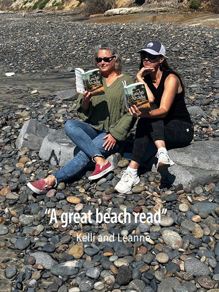 Two women sitting on a rocky beach, reading books. One woman wears a green sweater, blue jeans, and sunglasses; the other wears black clothing, a cap, and sunglasses.