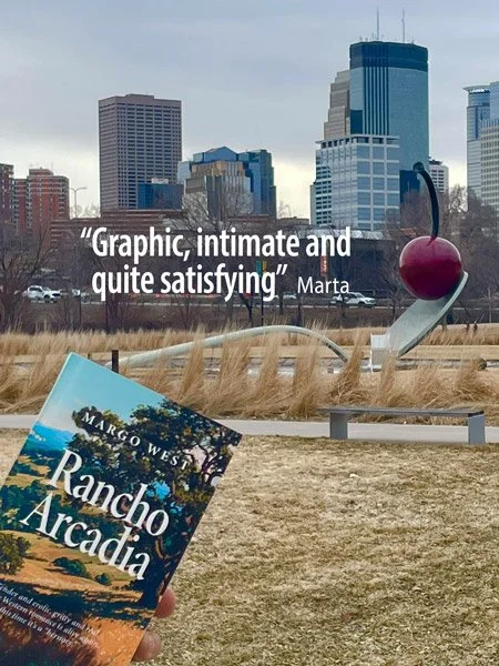 A cityscape with tall buildings in the background, a metal sculpture of a cherry on a spoon, a book titled "Rancho Arcadia" by Margo Wes, and a quote overlay that reads "Graphic, intimate and quite satisfying" by Marta.
