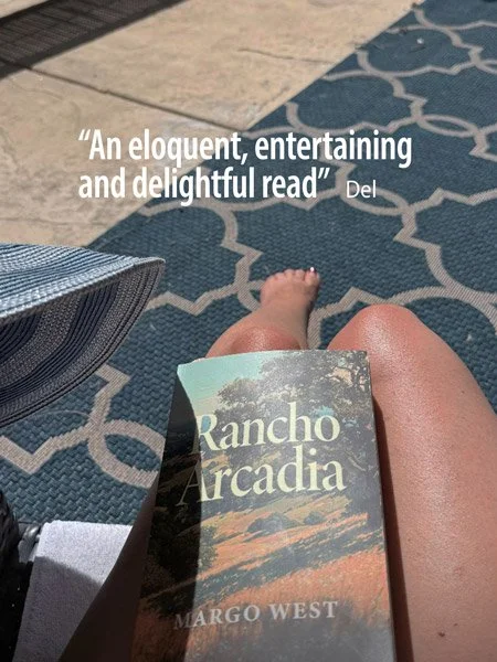 A person sitting outdoors with their legs extended, holding a book titled 'Rancho Arcadia' by Margo West. The background shows a patterned outdoor rug and a chair. A quote reading 'An eloquent, entertaining and delightful read' by Del is overlaid on the image.