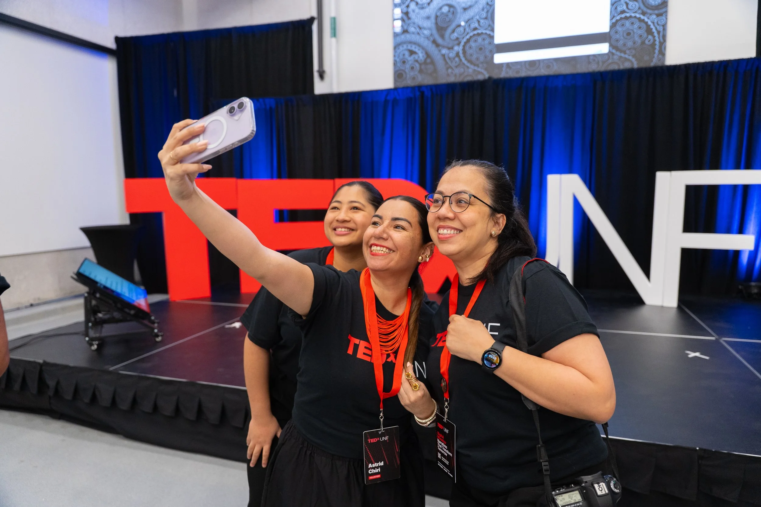 Three women taking a selfie at a TED event with large red and white TED letters in the background, standing on a stage with blue curtains.