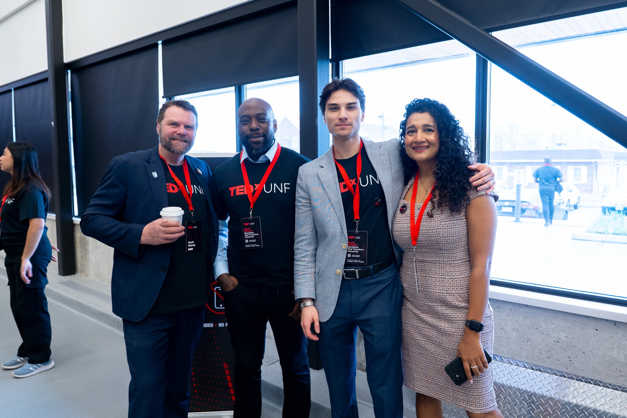 Group of four diverse people standing in front of glass windows at an indoor event, smiling and wearing conference badges, with two in professional attire and two in casual clothing.