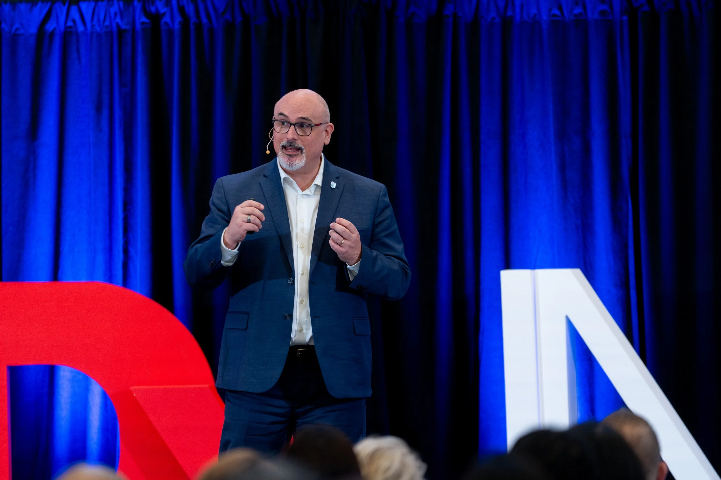 A man in a suit and glasses speaking at an event with large red, white, and blue balloons and a blue curtain backdrop.