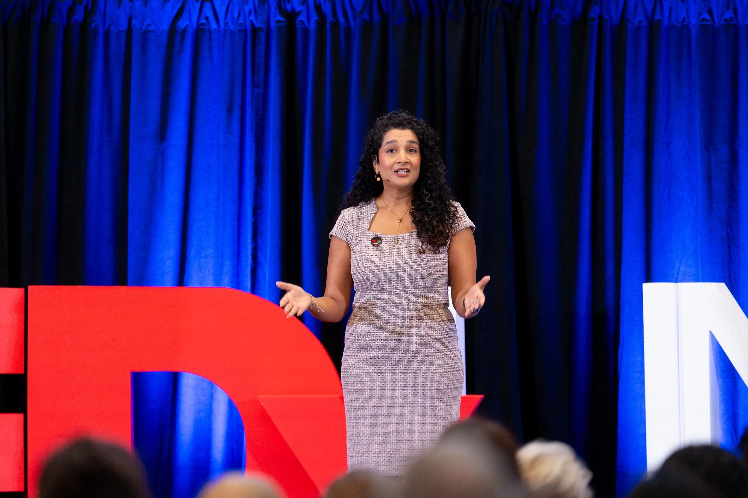 A woman with curly dark hair is speaking on stage during a presentation. She is wearing a beige patterned dress and standing in front of a blue curtain with large red, white, and blue symbols or display elements.