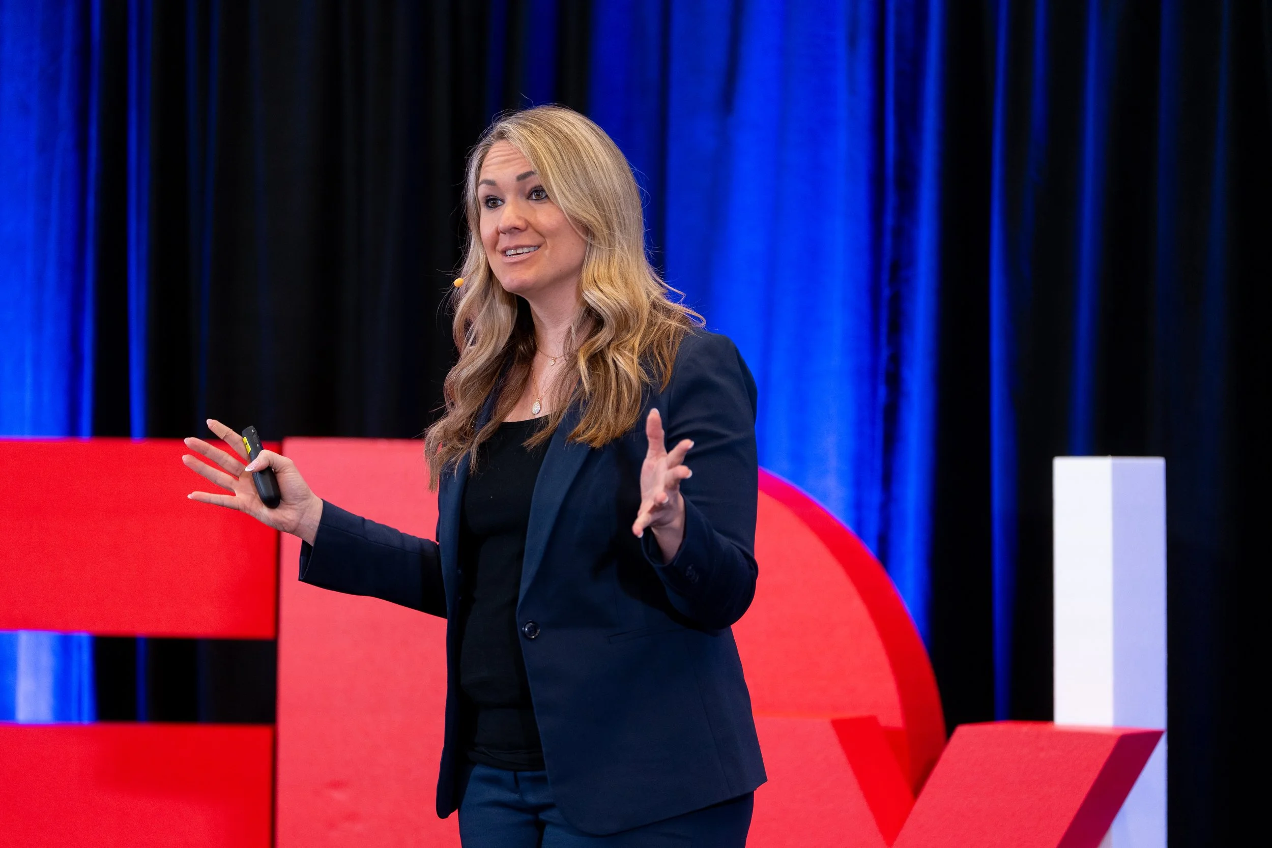 A woman with long blonde hair wearing a dark blazer and black shirt is speaking on stage, gesturing with her right hand while holding a remote in her left hand. The background features red, black, and blue curtains.