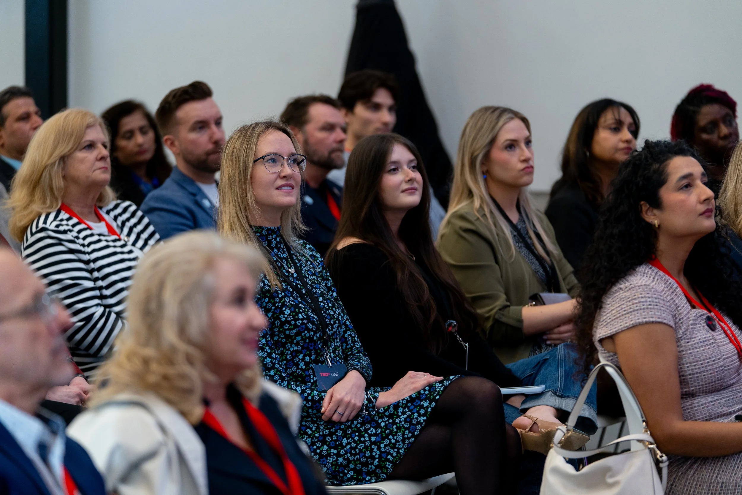 Audience members sitting and listening at an indoor event, some with focused or attentive expressions, including a woman with glasses and a blue patterned dress.