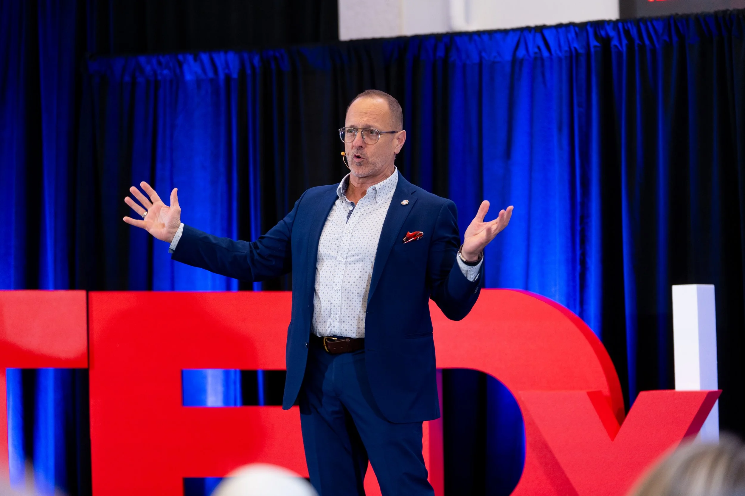 A man in a dark suit with a white patterned shirt and glasses speaks on a stage with blue curtains in the background. He is gesturing with his hands during a presentation or speech, standing behind a red podium.
