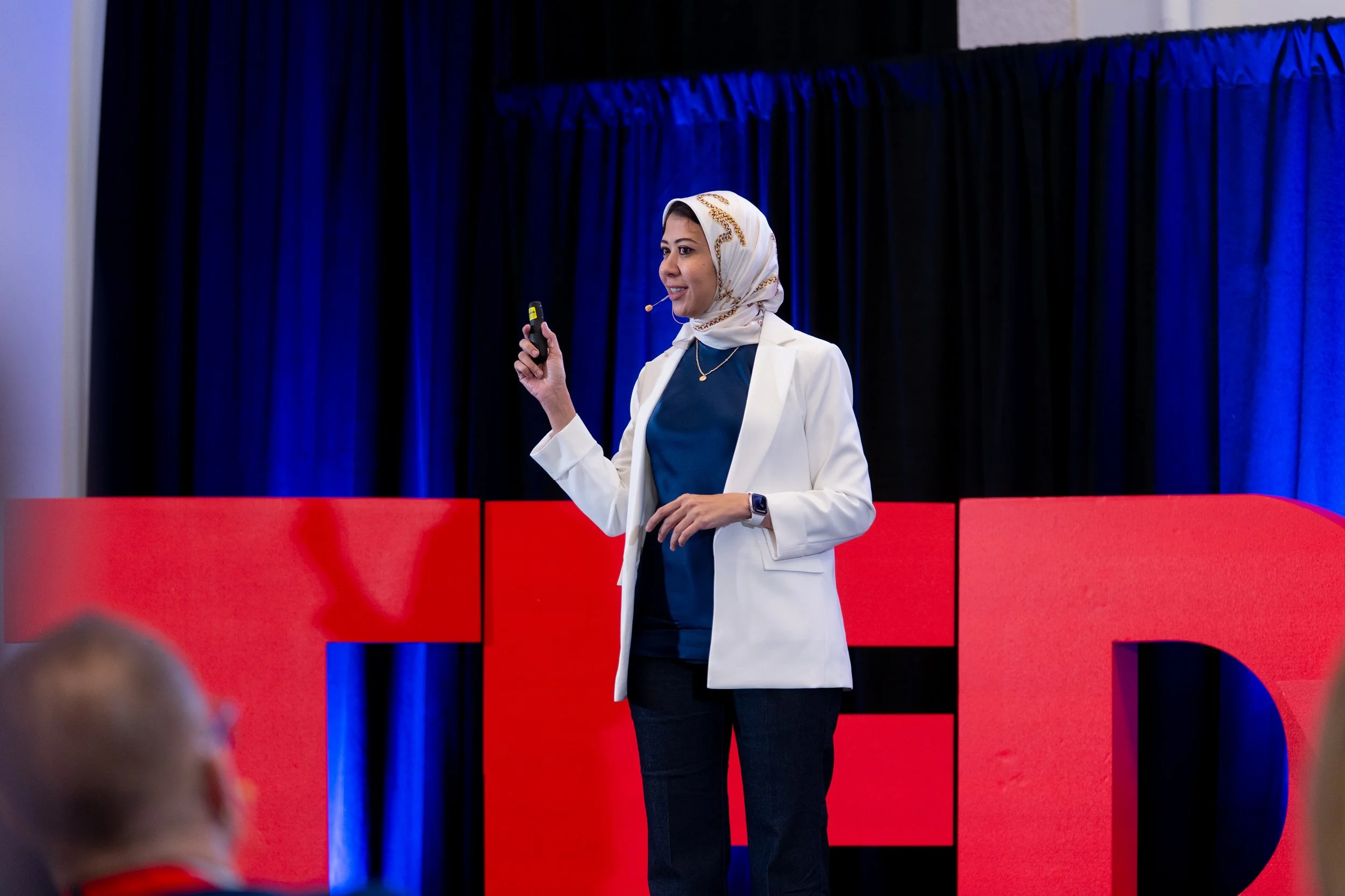 A woman wearing a white blazer and black pants giving a presentation with a microphone headset, standing in front of red and black panels and blue curtains.