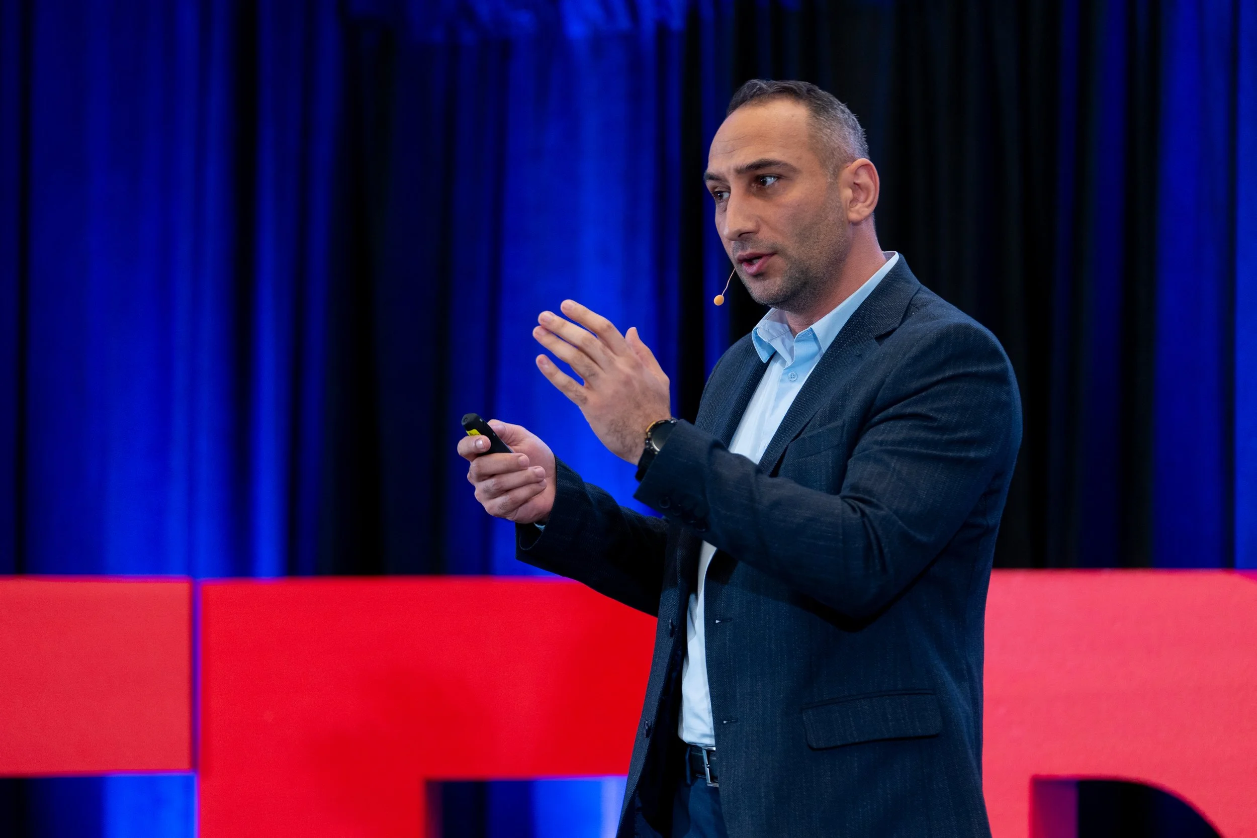 A man in a dark suit and light blue shirt speaking on stage with a microphone headset, holding a remote control device, against a background of blue curtains and red stage floor.