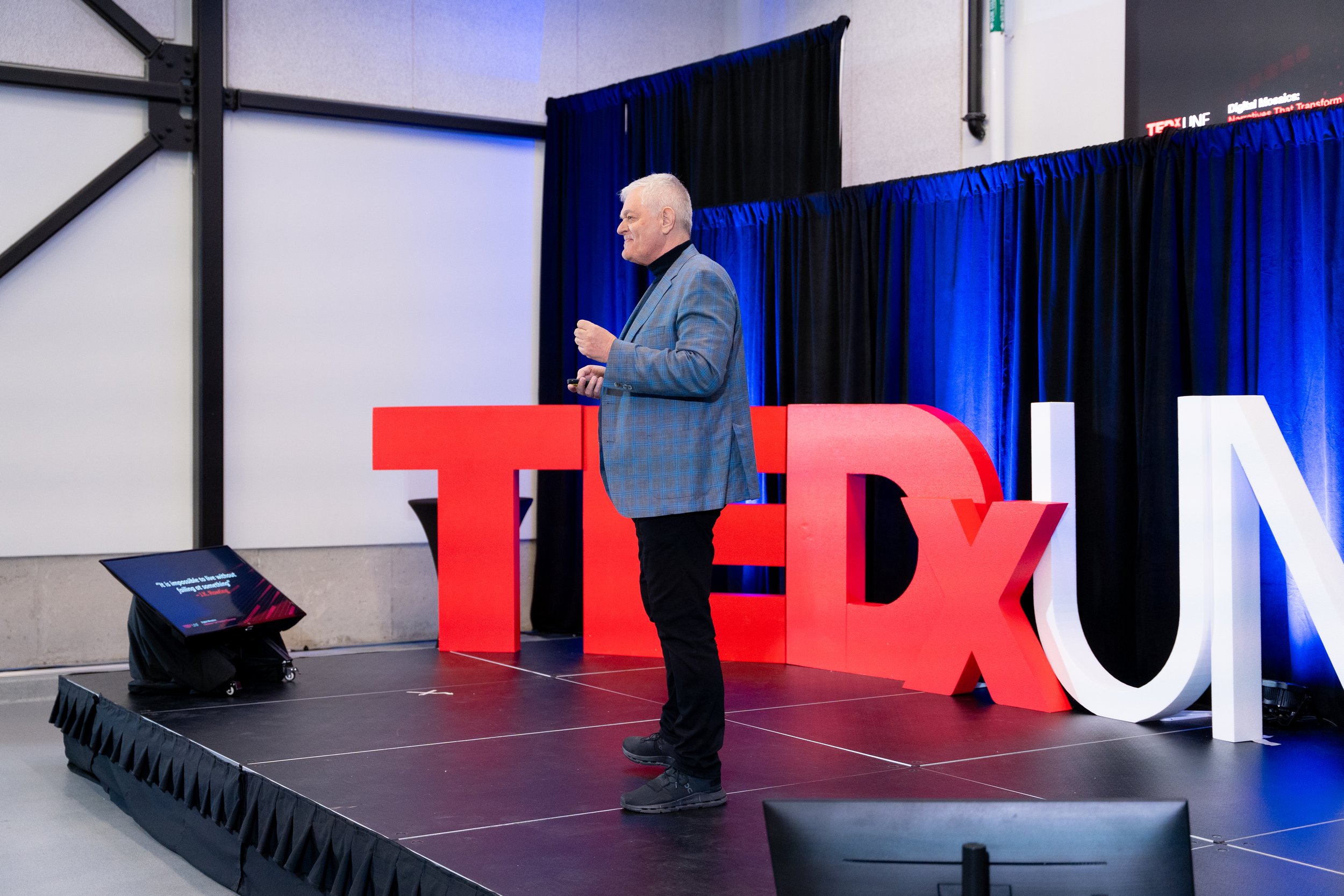 A man with gray hair wearing a gray blazer with a plaid pattern giving a presentation on a stage with large red and white letters spelling TEDx. Black curtains are behind him and a screen displaying TEDx branding is visible.