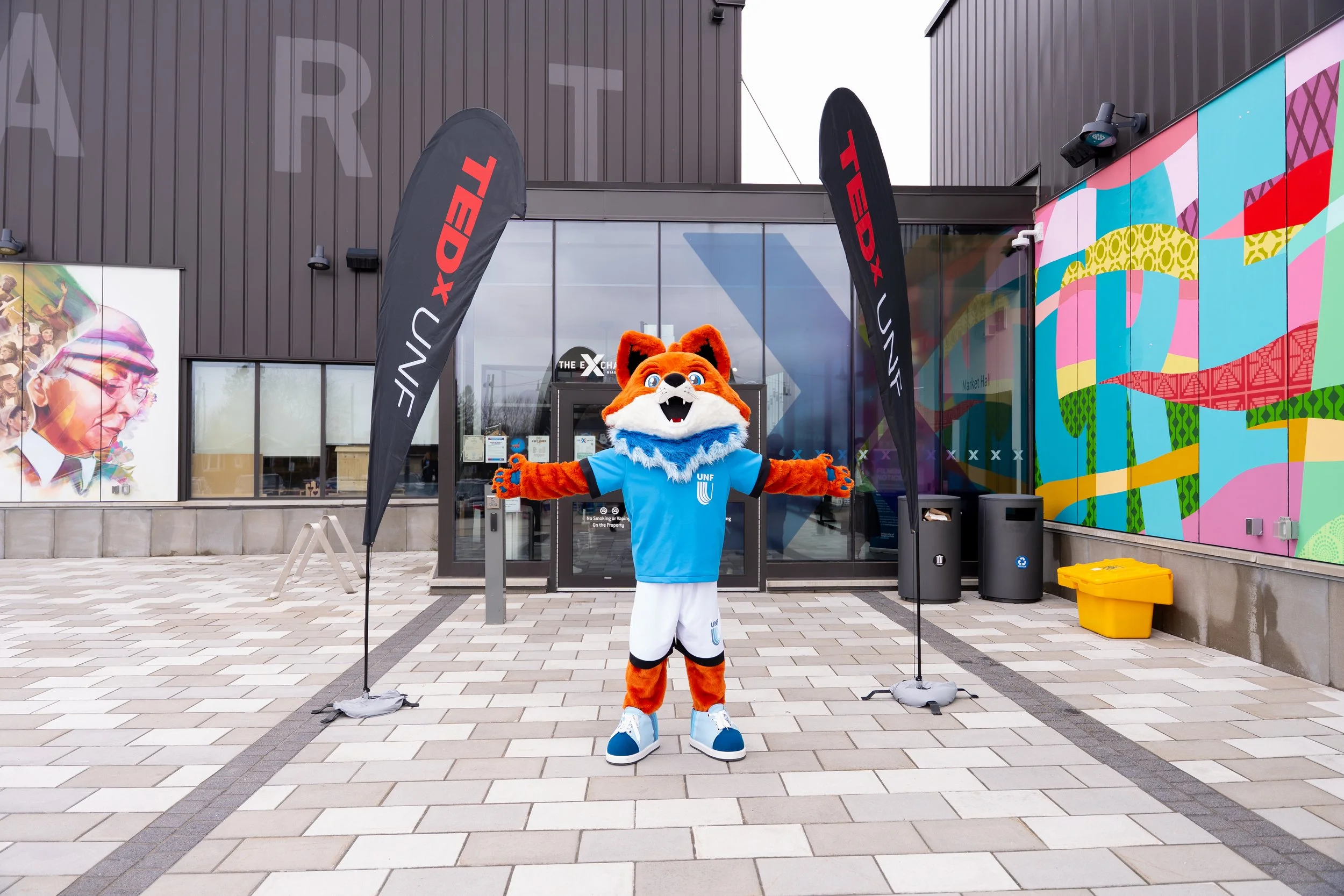 A mascot dressed as a fox wearing a blue UNF t-shirt with arms outstretched stands on a tiled outdoor area in front of a modern building. Behind the mascot, there are two black flags with red and white text that say 'TEDx'. Colorful abstract murals a