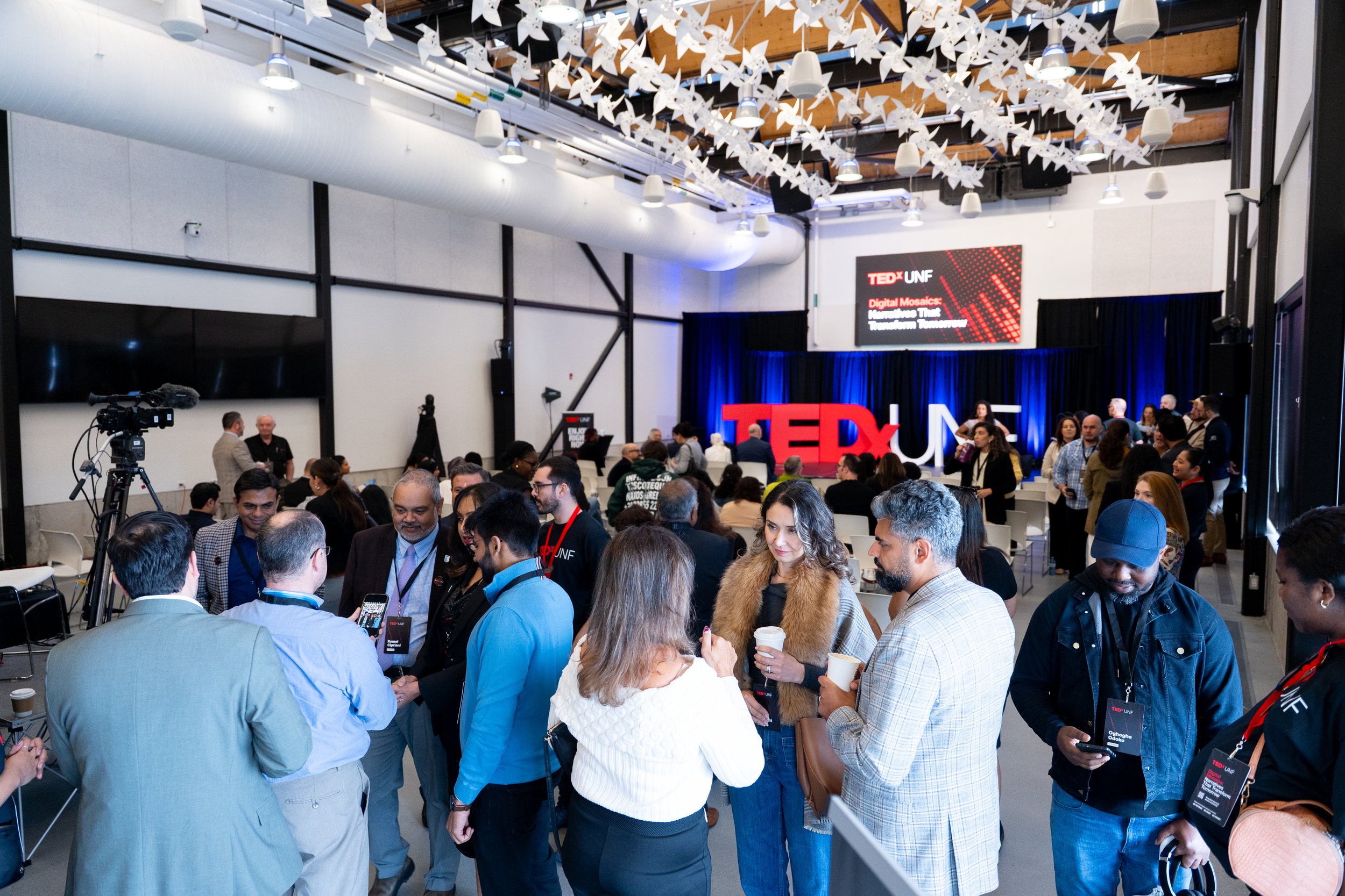 A crowded indoor event space with people gathered around, engaging in conversations, with a large screen displaying 'TEDx UNF' and the theme 'Digital Mosaics: Navigating Threads, Transforming Tomorrow' in the background. Ceiling decorations include numerous white pinwheels.