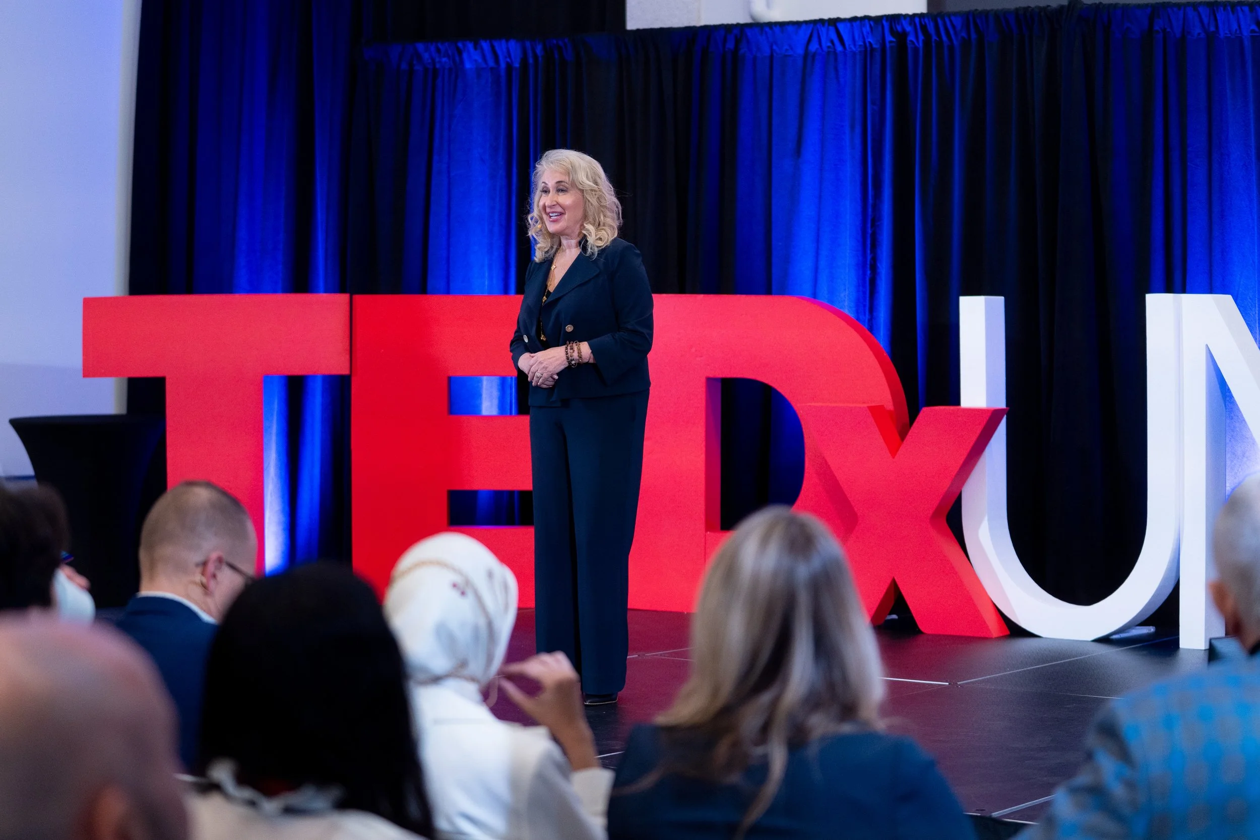 A woman speaking on stage at a TEDx event with large red and white TEDx letters behind her, blue curtains in the background, and an audience in the foreground.