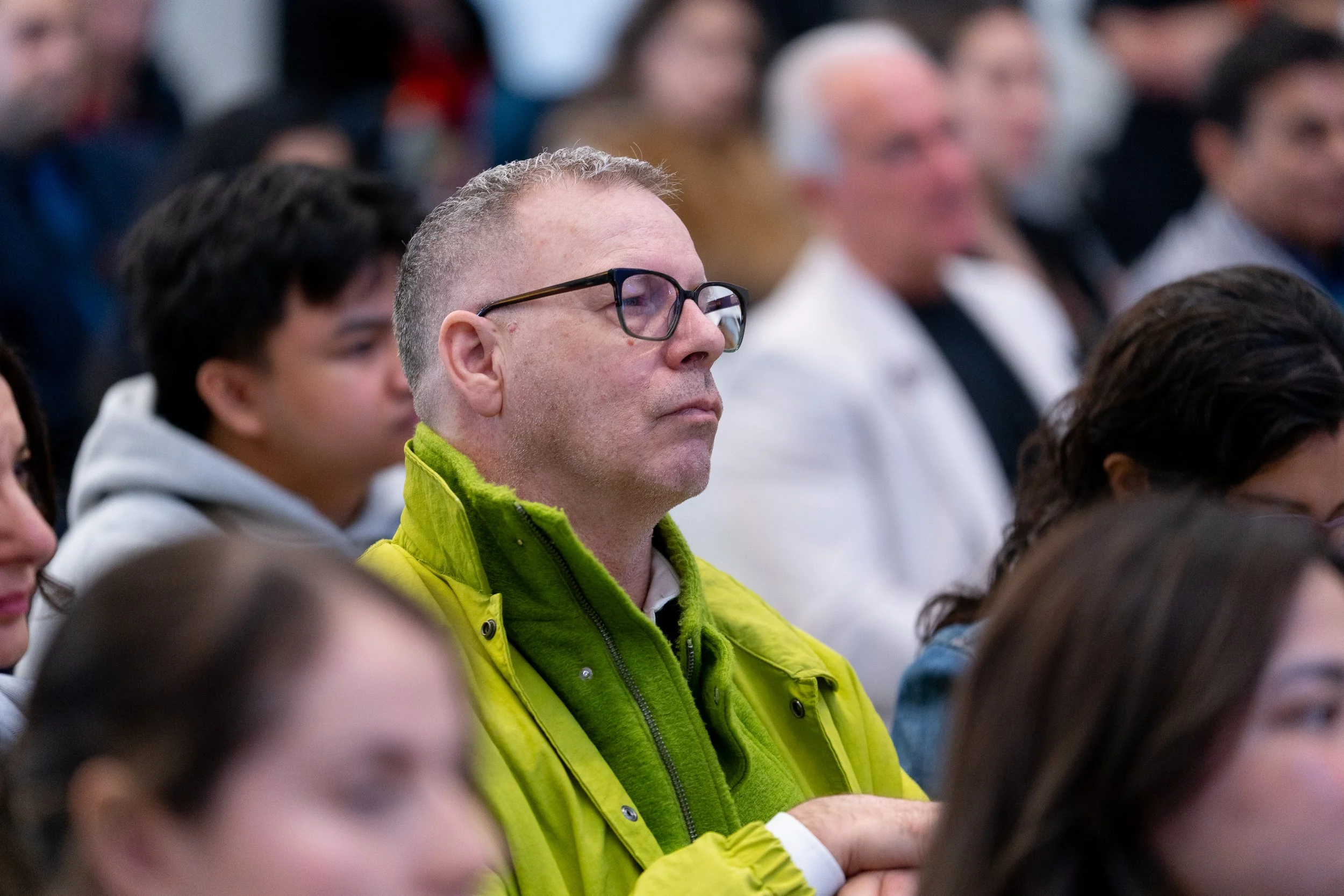 A man with glasses wearing a bright green jacket, sitting among a crowd, listening attentively.