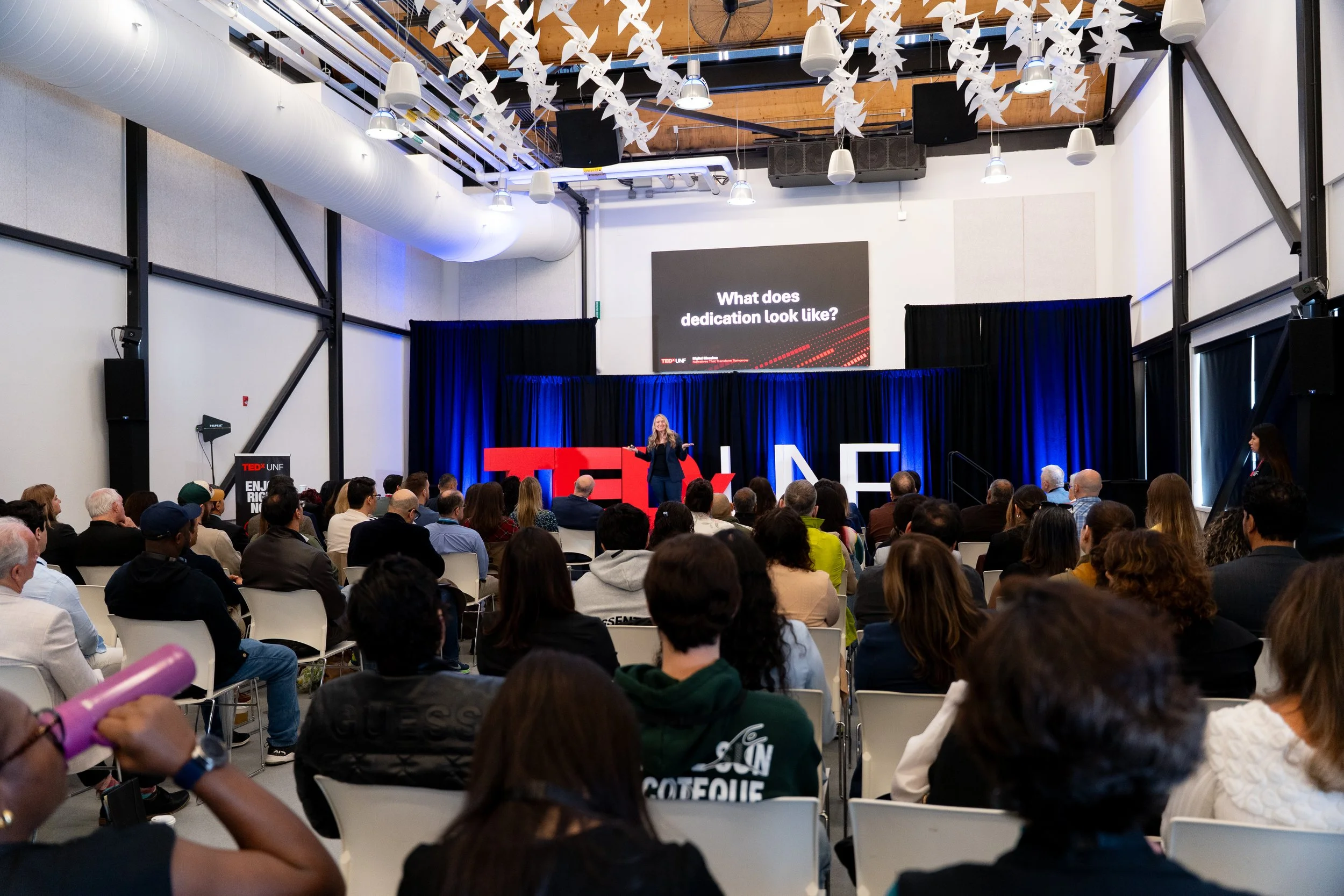 A woman giving a presentation at a TEDx event with a large audience present. There is a screen behind her with the question, "What does dedication look like?" The setting appears to be a modern, industrial-style hall with high ceilings and hanging de