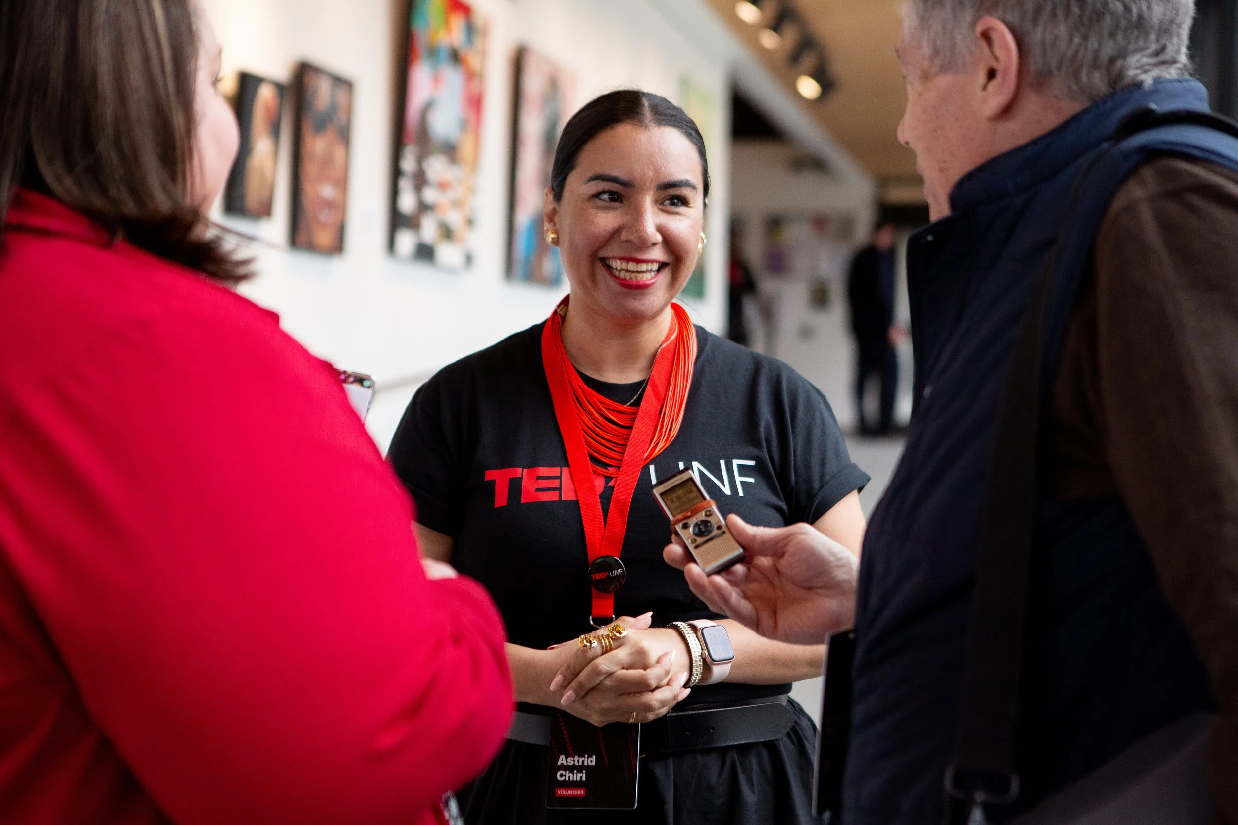 A woman smiling and talking to two people in an indoor setting, with artwork on the wall behind her. She is wearing a black T-shirt with the text 'TED UNC' and a red lanyard with a name badge. The woman has dark hair pulled back and is holding a smal