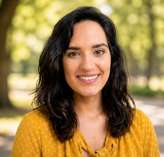 A woman with dark, wavy hair smiling outdoors, wearing a yellow blouse with a dotted pattern, in a park with trees in the background.