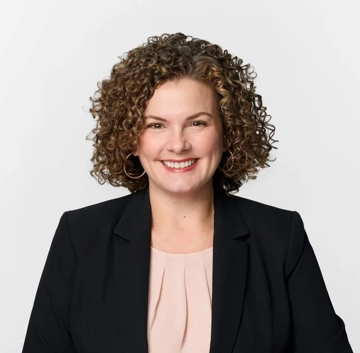 Professional woman with curly brown hair smiling, wearing a black blazer and light pink blouse against a plain white background.