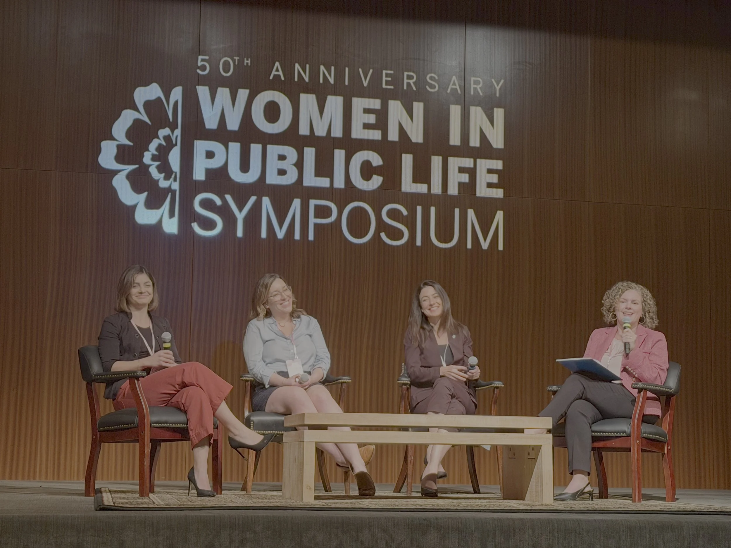 Four women seated on stage during the 50th anniversary of Women in Public Life Symposium, smiling, with three holding microphones, against a wood-paneled background with the event logo.
