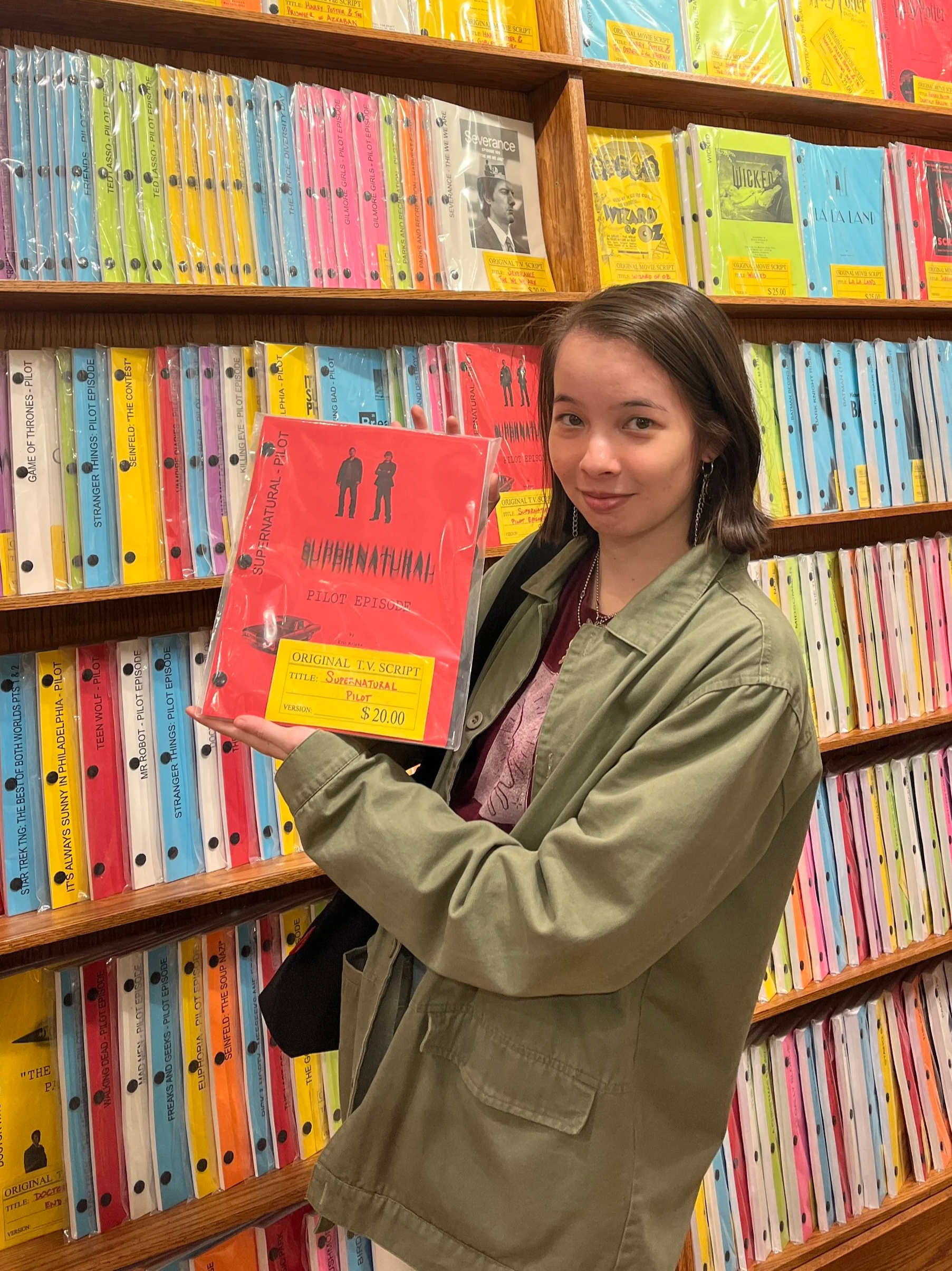A young woman with shoulder-length brown hair and hoop earrings standing in front of a bookshelf filled with colorful scripts, holding a red script titled 'Supernatural Pilot Episode' at an indoor location.