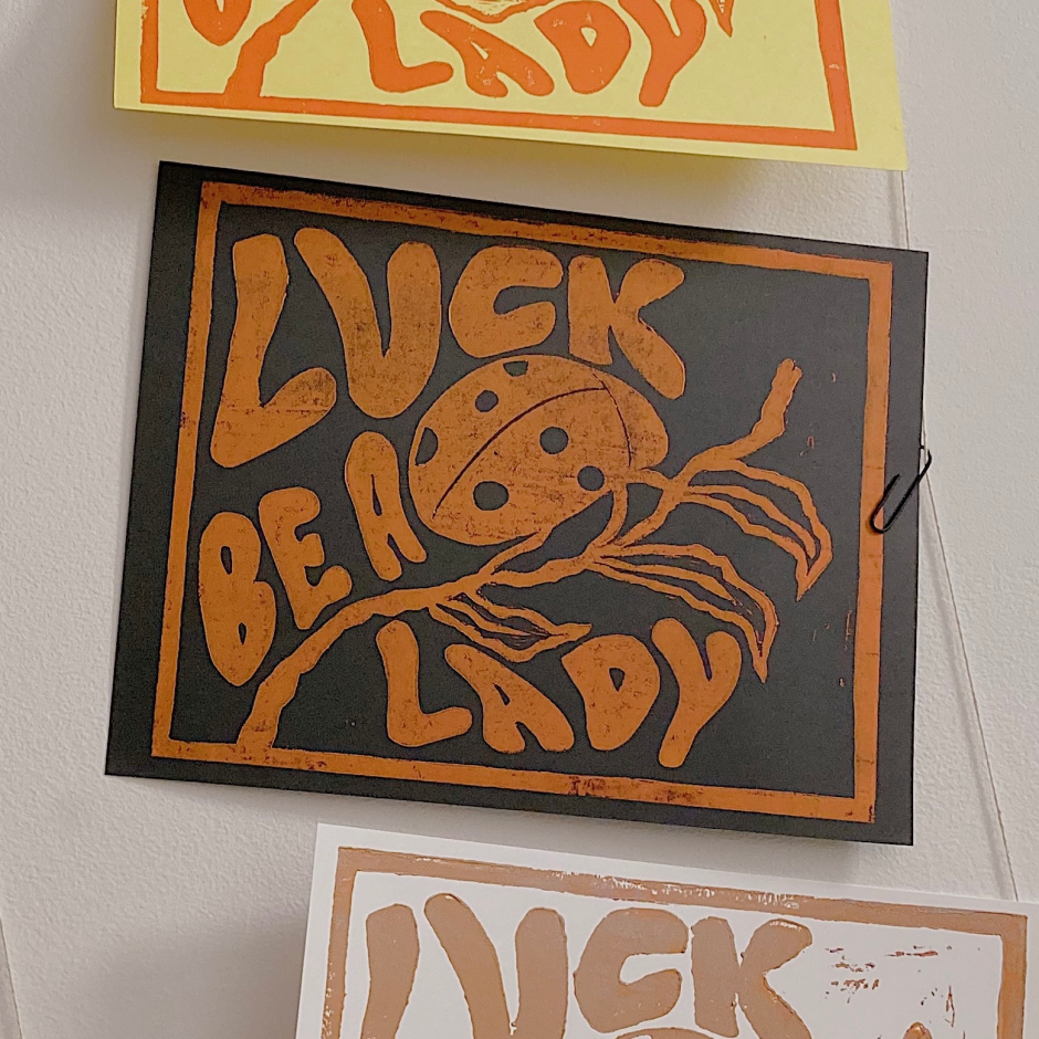 A black paper with orange text and design that says 'LUCK BE A LADY' with a ladybug illustration and decorative lines. It is attached to a wall with a paperclip.