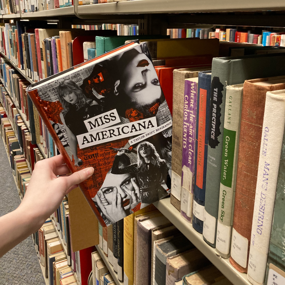 Hand holding a book titled 'Miss Americana' by Taylor Swift in a library aisle with shelves of books.