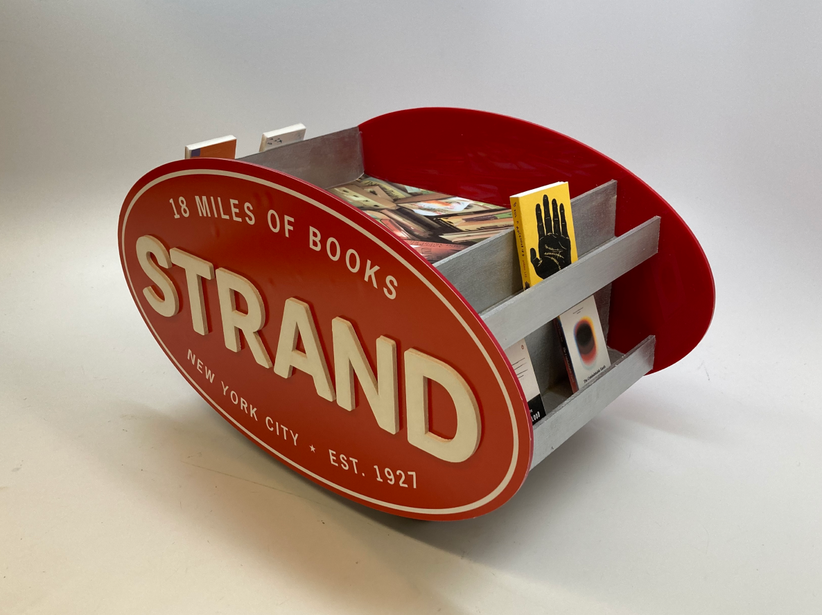 A red and gray wooden display bin with a sign reading 'STRAND.' The bin contains various books.