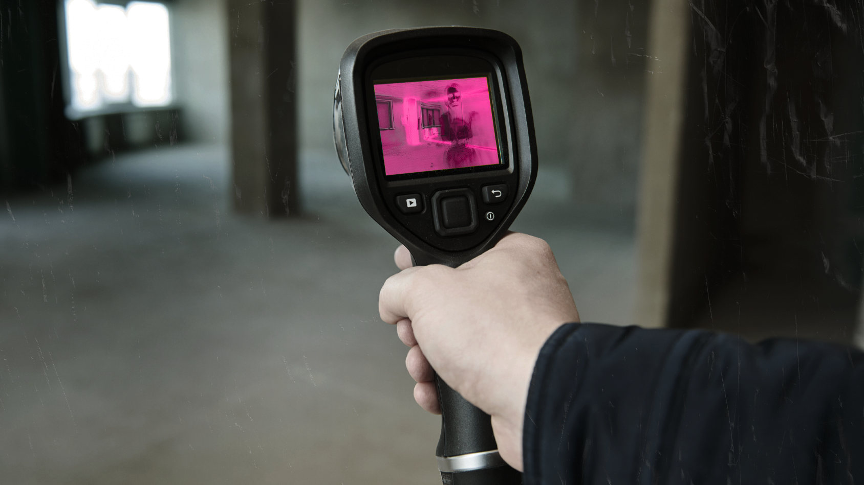 A person holding a thermal imaging camera displaying a pink and black thermal image of a room and a person inside.