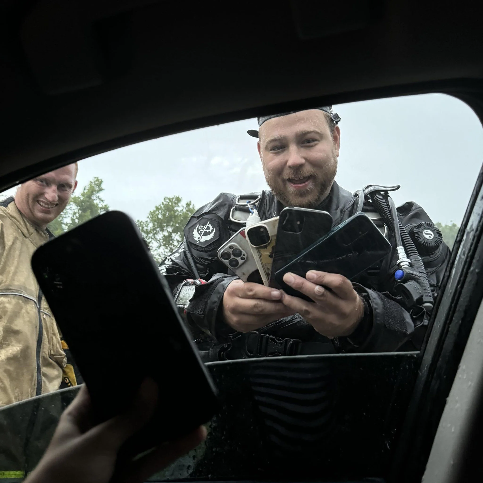 A man in motorcycle gear and a woman are seen through the windshield of a vehicle, smiling and looking at the camera during daytime with a cloudy sky and some trees in the background.