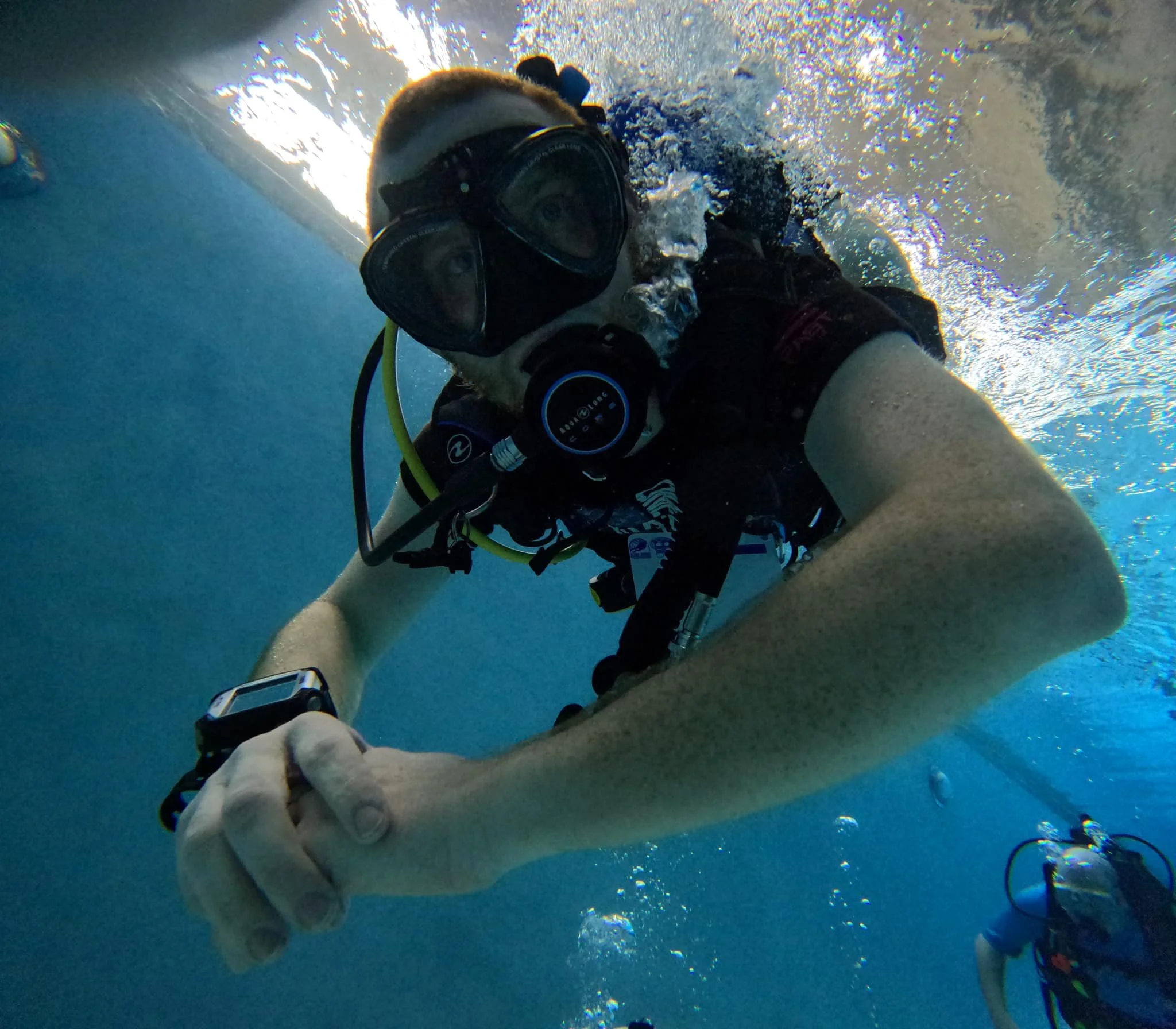 A person taking a selfie underwater while scuba diving, with a dog looking up at the camera. The diver is wearing a mask, snorkel, and wetsuit, and bubbles are visible around them.