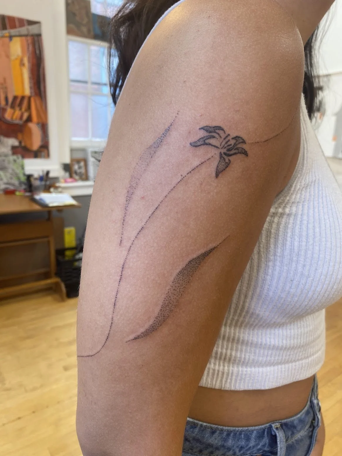A person's upper arm with a fresh tattoo of a flower and abstract designs, in a room with wooden floors, a desk, and shelves.