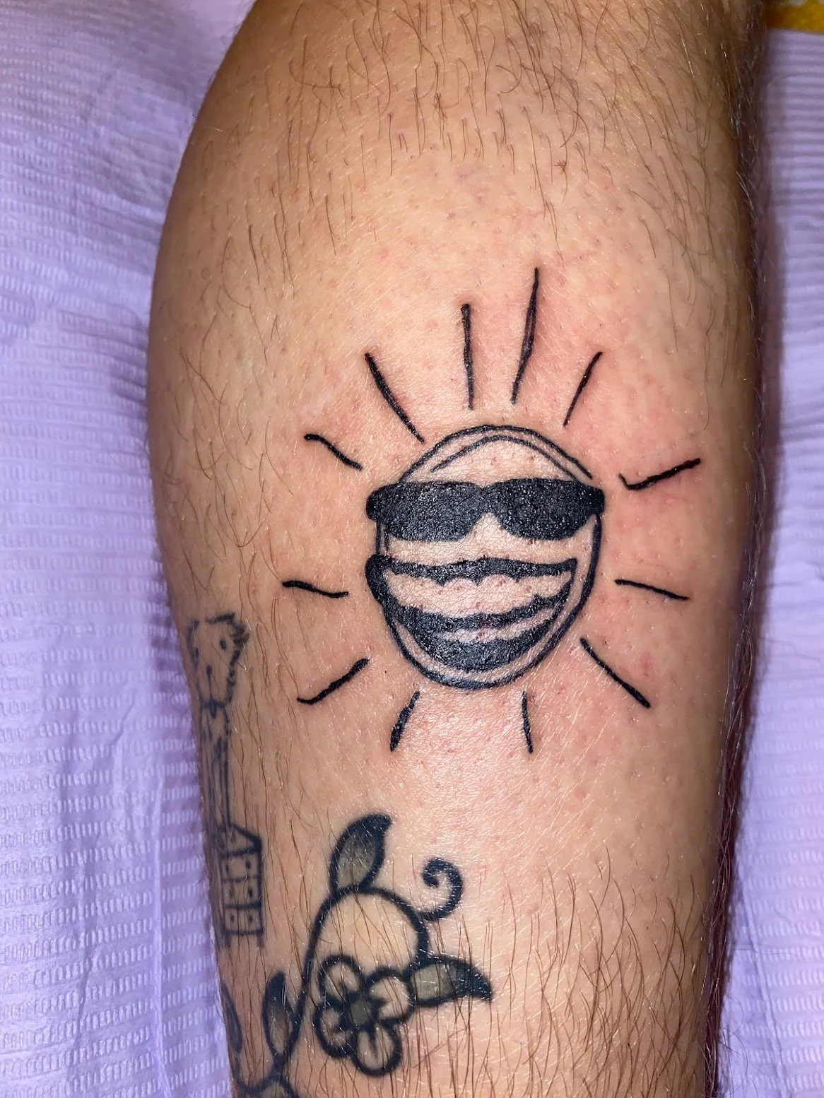 Tattoo of a smiling sun wearing sunglasses, surrounded by rays, on a person's arm.