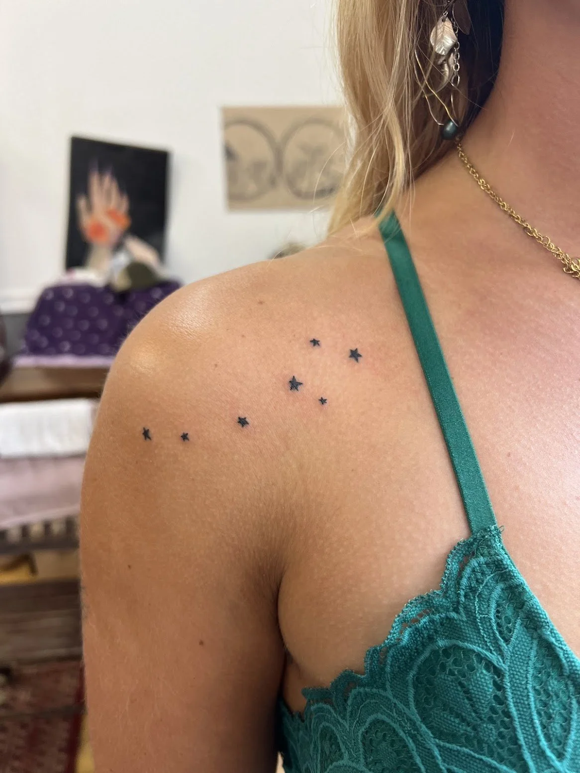 Close-up of a woman's shoulder and upper chest with a tattoo of small black stars, the little dipper constellation. She is wearing a teal lace top, a gold chain necklace, and an earring with beads. The background shows blurry art and a purple bag.