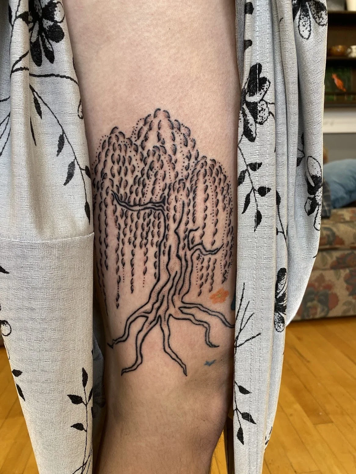 A tattoo of a willow tree with branches and roots on a person's thigh, with vertical lines representing rain above the tree.