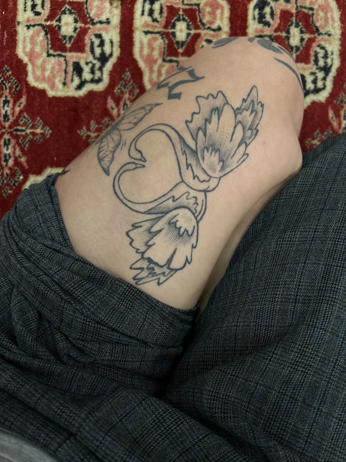 A person's upper thigh with a tattoo of two swans nose to nose forming a heart. The person is wearing dark checkered clothing and is sitting on a patterned red, black, and cream rug.