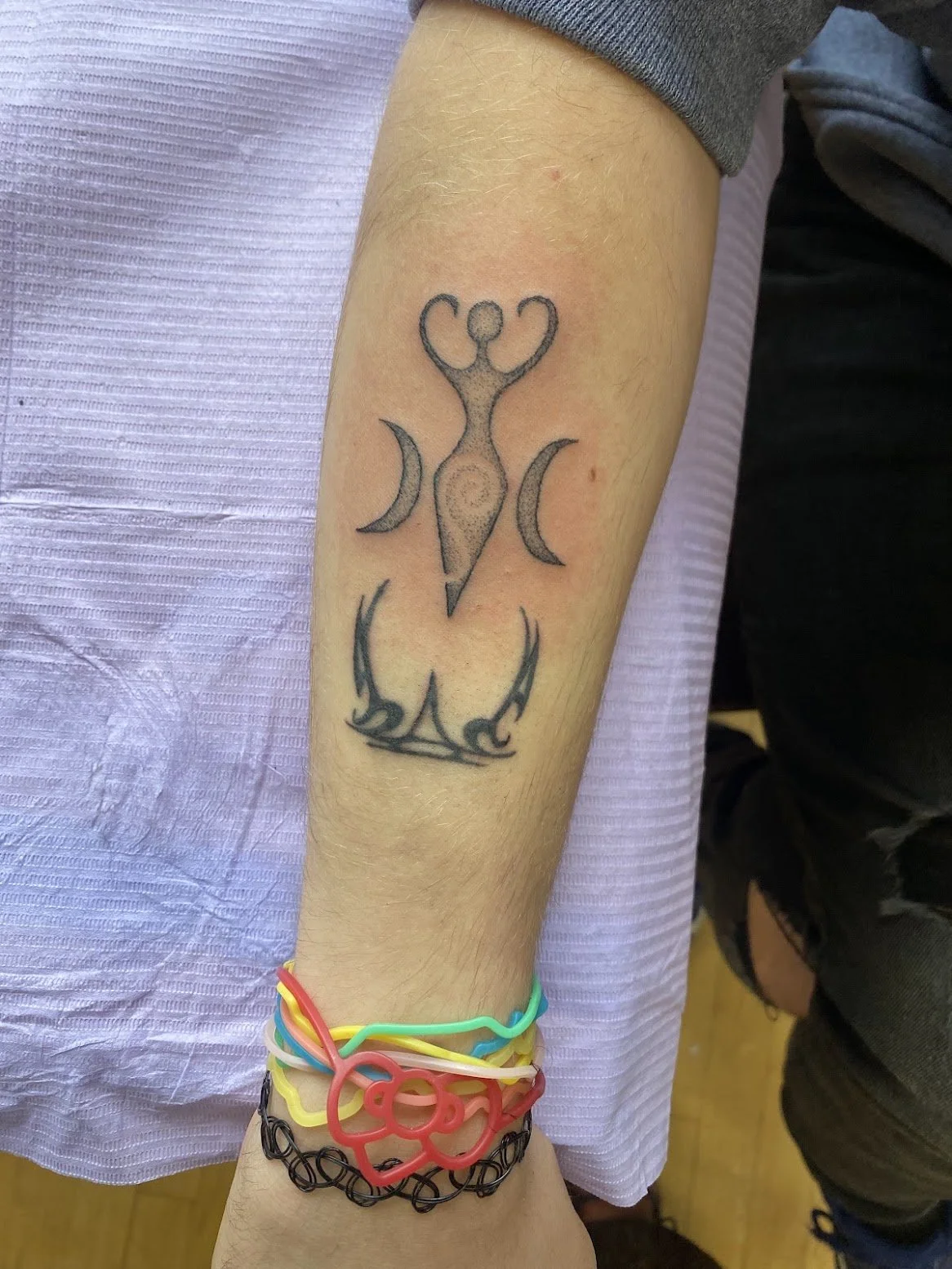 Tattoo of a stylized female figure with raised arms, flanked by crescent moons on each side, the divine feminine, and a semi-circular element with flames or spikes below, on a person's forearm.