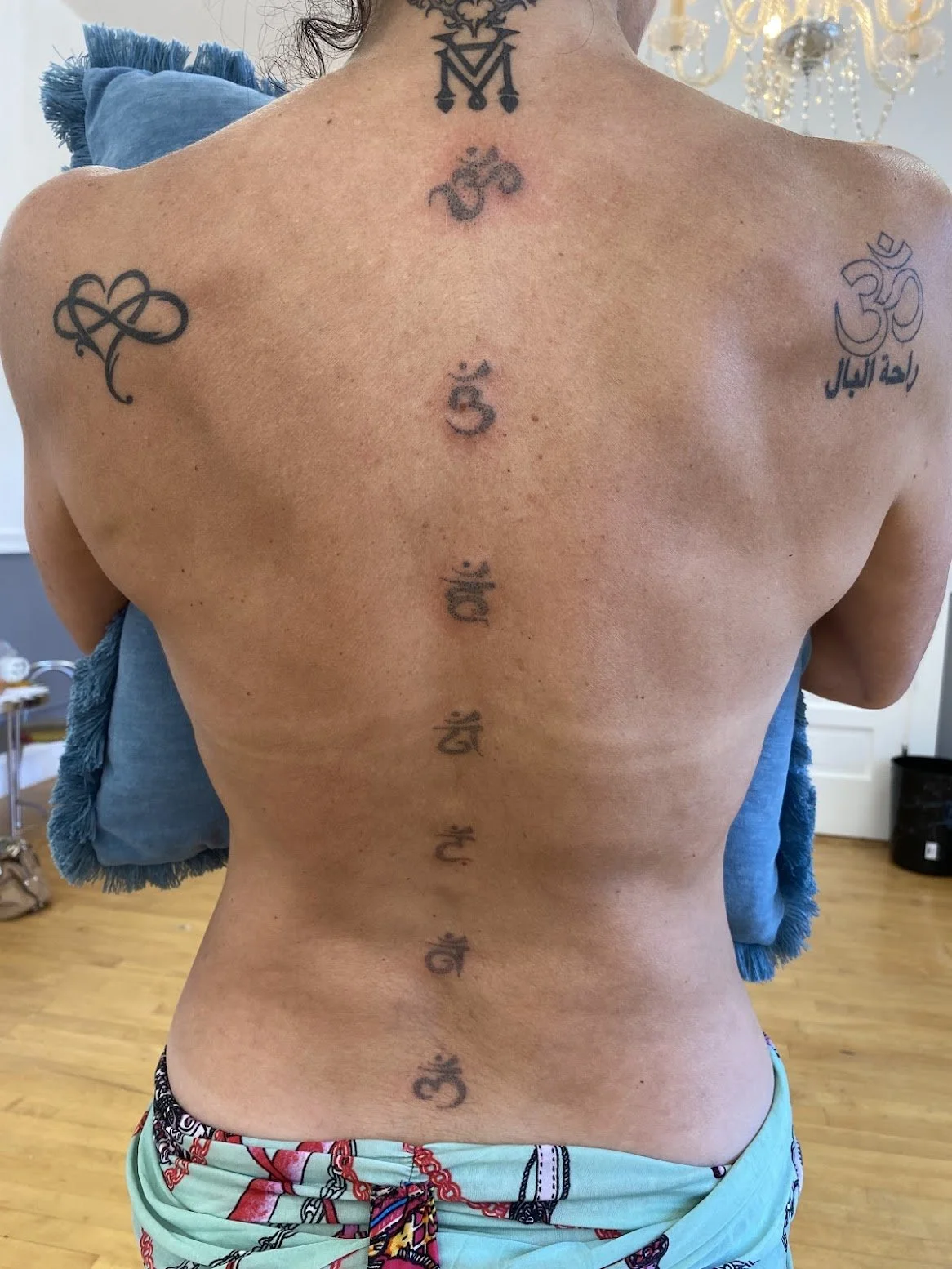 Person's back with tattoos, including seven chakra symbols down the spine and various symbols on shoulders and neck, in a room with blue pillows and a chandelier.