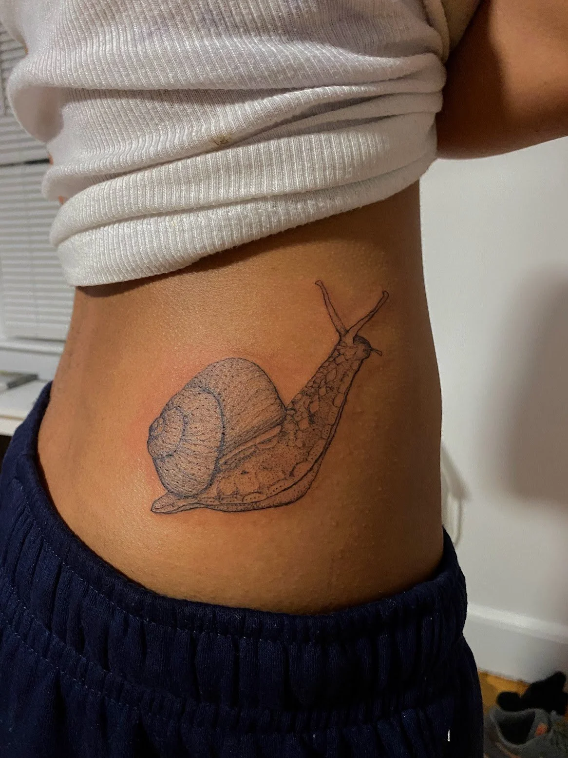 Tattoo of a snail on a person's side, with a detailed outline and shading, above the waistband of dark blue shorts and below a white ribbed shirt.