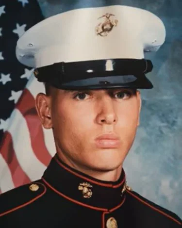 Young Marine in dress blues uniform and white cap standing in front of an American flag.