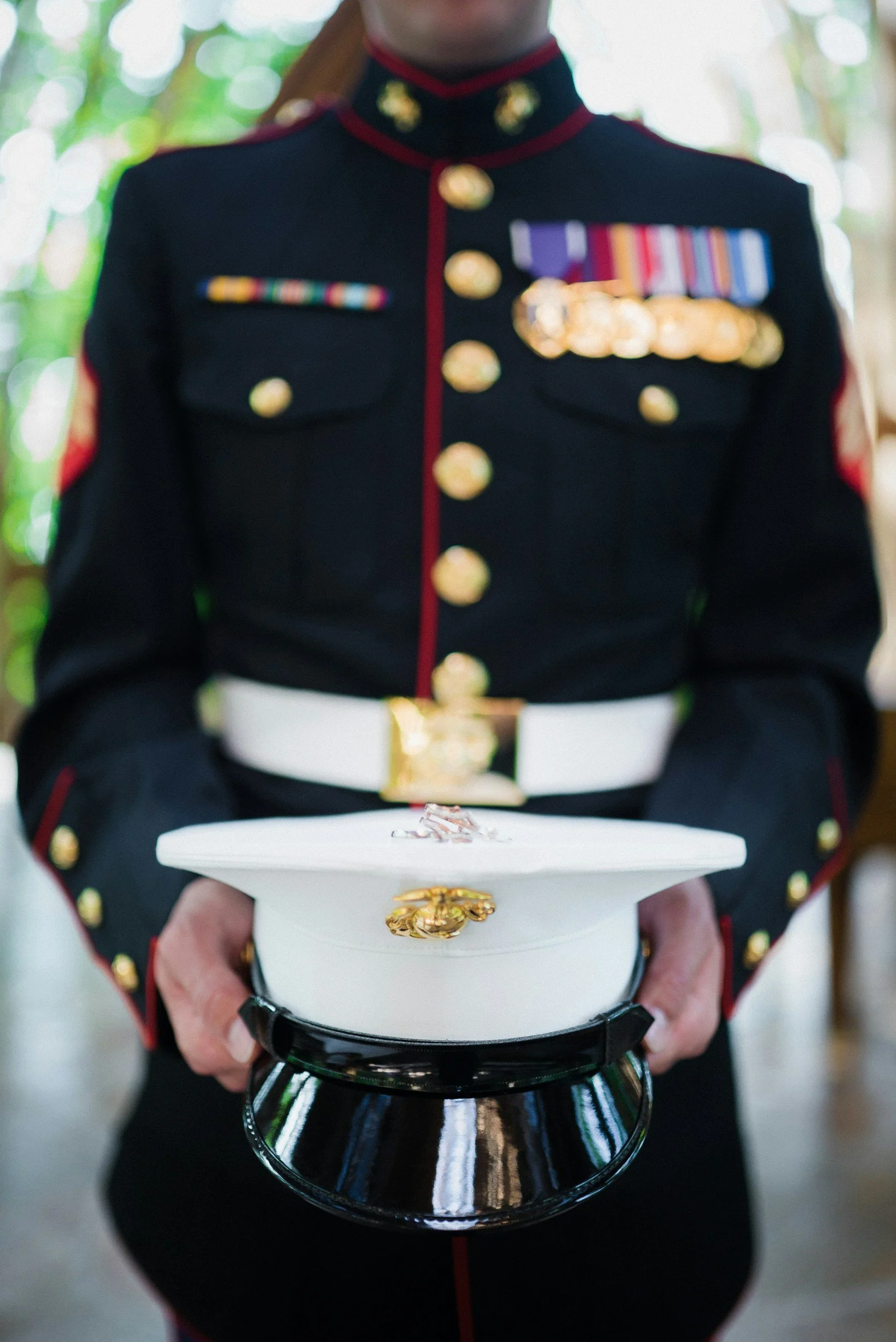 A person dressed in a United States Marine Corps dress uniform holding a white hat with a gold emblem in front of them.