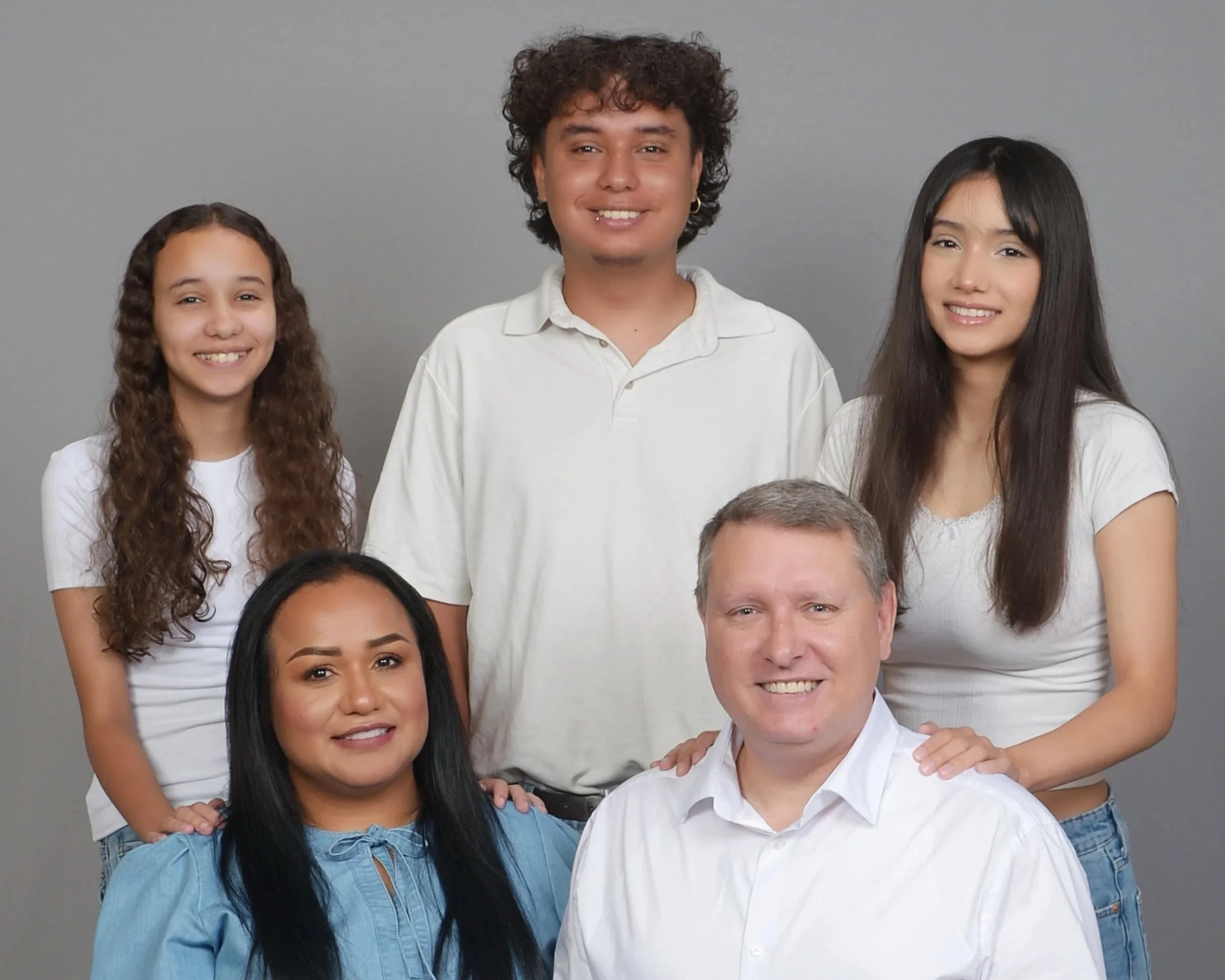 A diverse family of six people, including three men and three women, standing against a gray background, smiling and looking at the camera.